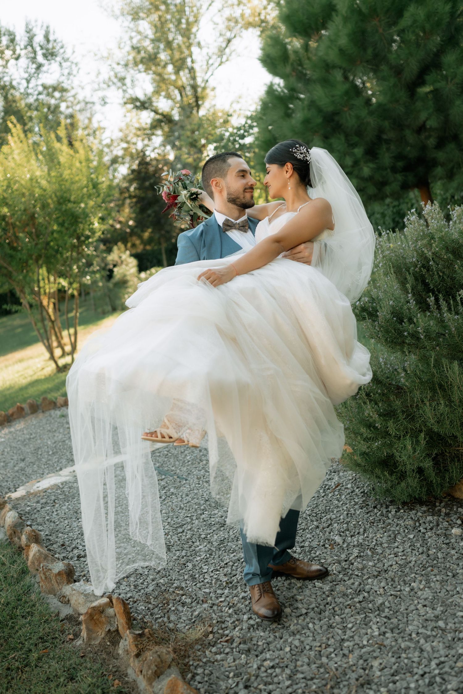 A romantic wedding couple shares a playful dip pose on a gravel path surrounded by greenery in golden afternoon light.