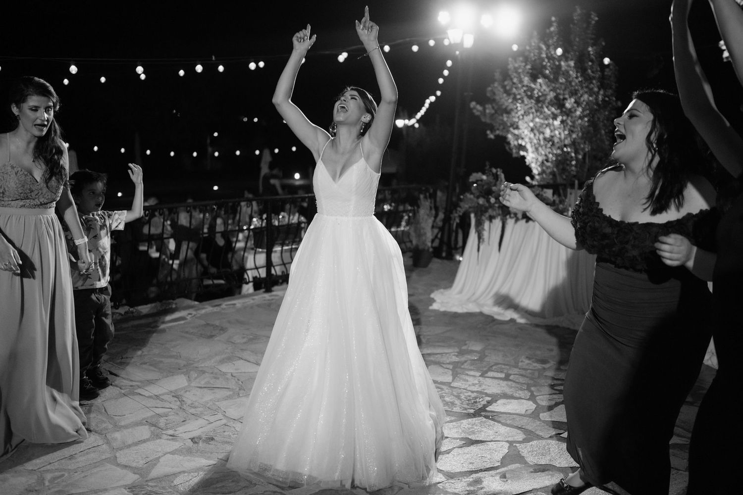 A joyful celebration on a stone dance floor at an outdoor evening wedding reception with string lights overhead.