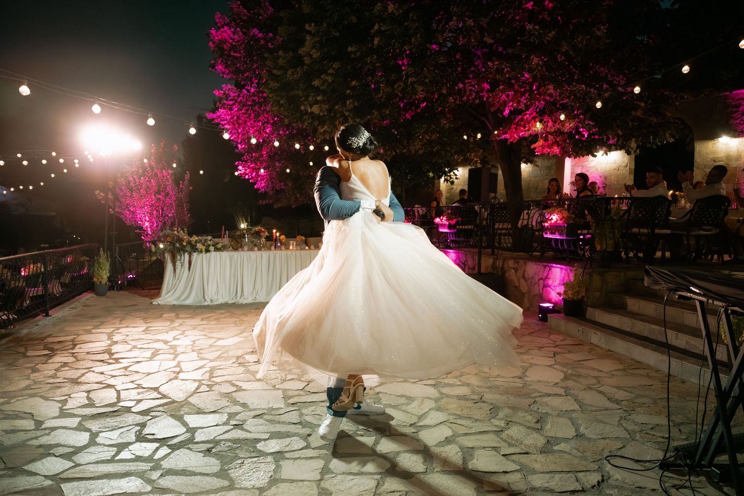 A couple shares their first dance at an outdoor wedding reception with pink uplighting and string lights illuminating the stone patio.