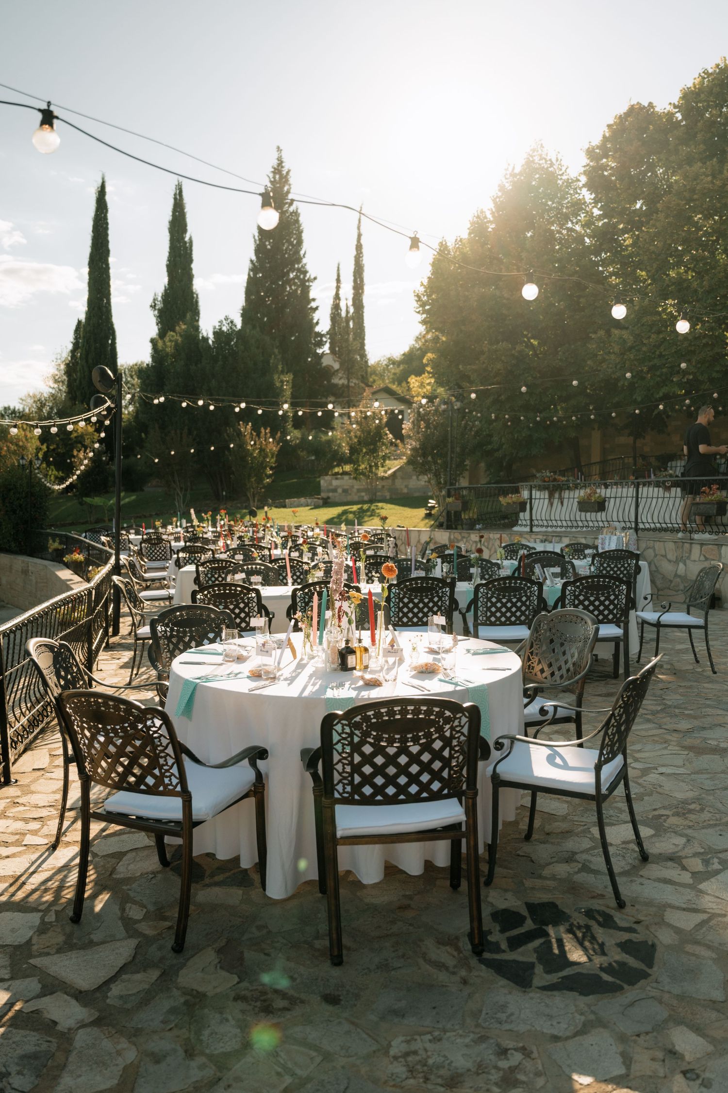 Outdoor wedding reception setup with round tables and metal chairs on stone patio under string lights at sunset.
