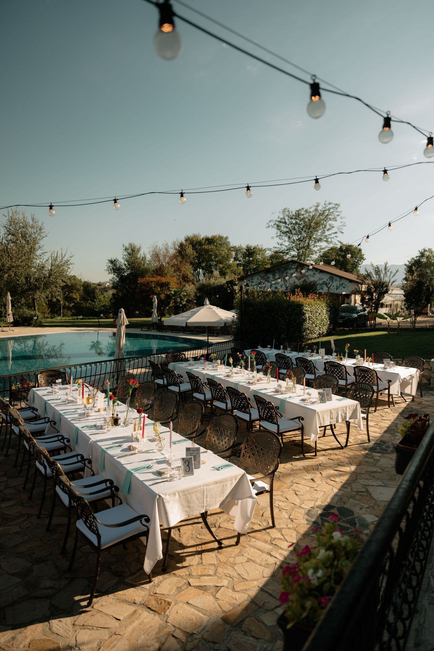 Elegant outdoor wedding reception setup with long tables and string lights by a swimming pool at sunset.