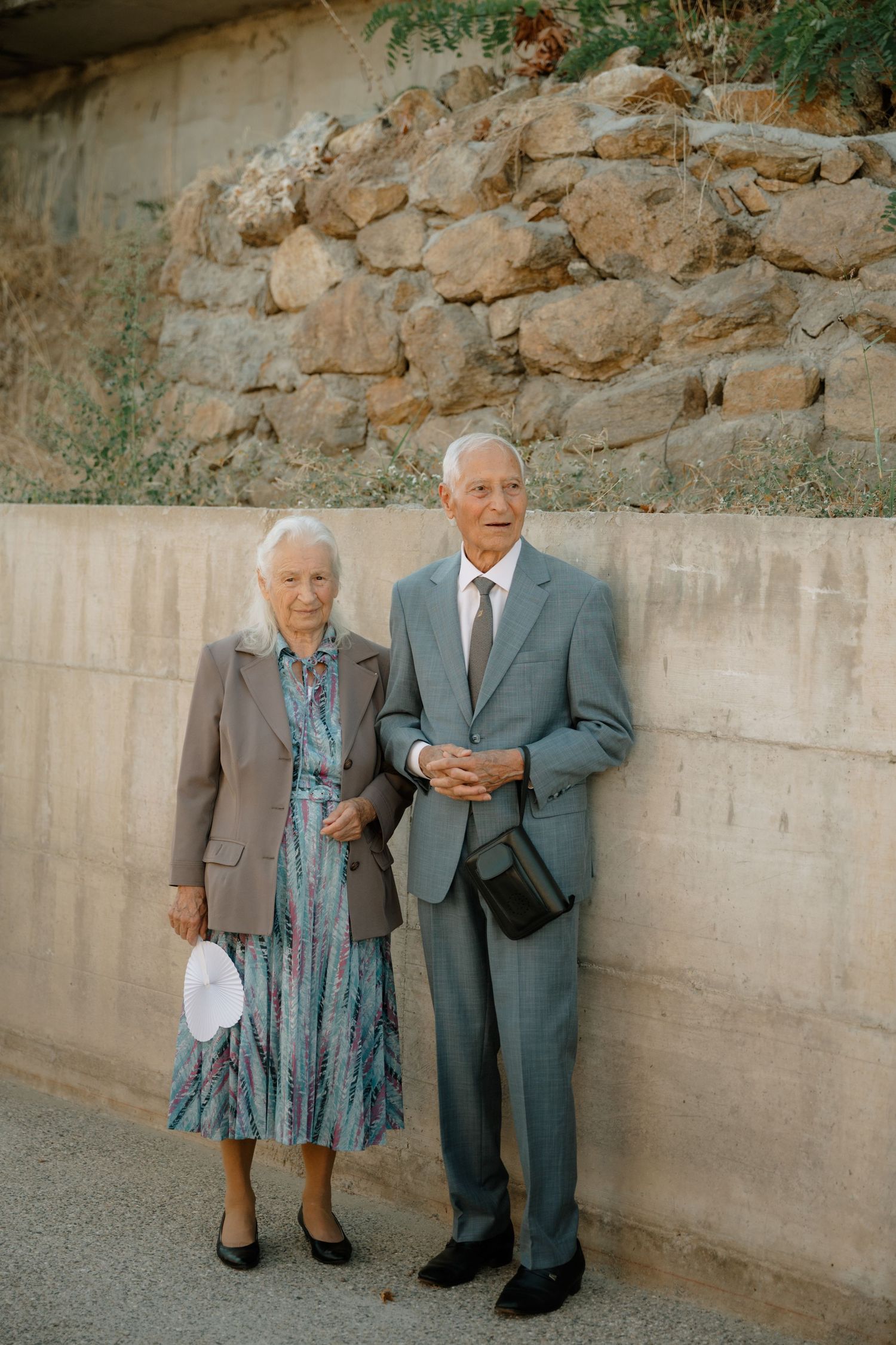 An elderly couple stands together against a stone wall, with the person in a suit gesturing while talking.