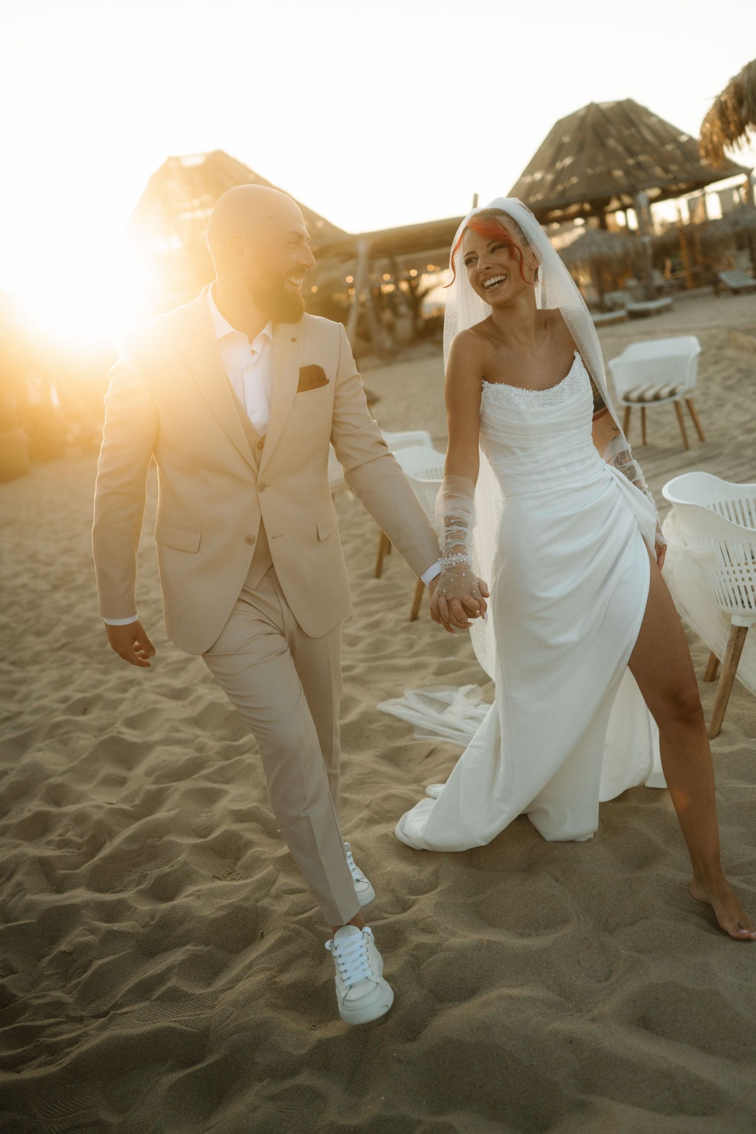 Newlyweds walking barefoot together on sandy beach at sunset with bride's white dress flowing in the breeze.
