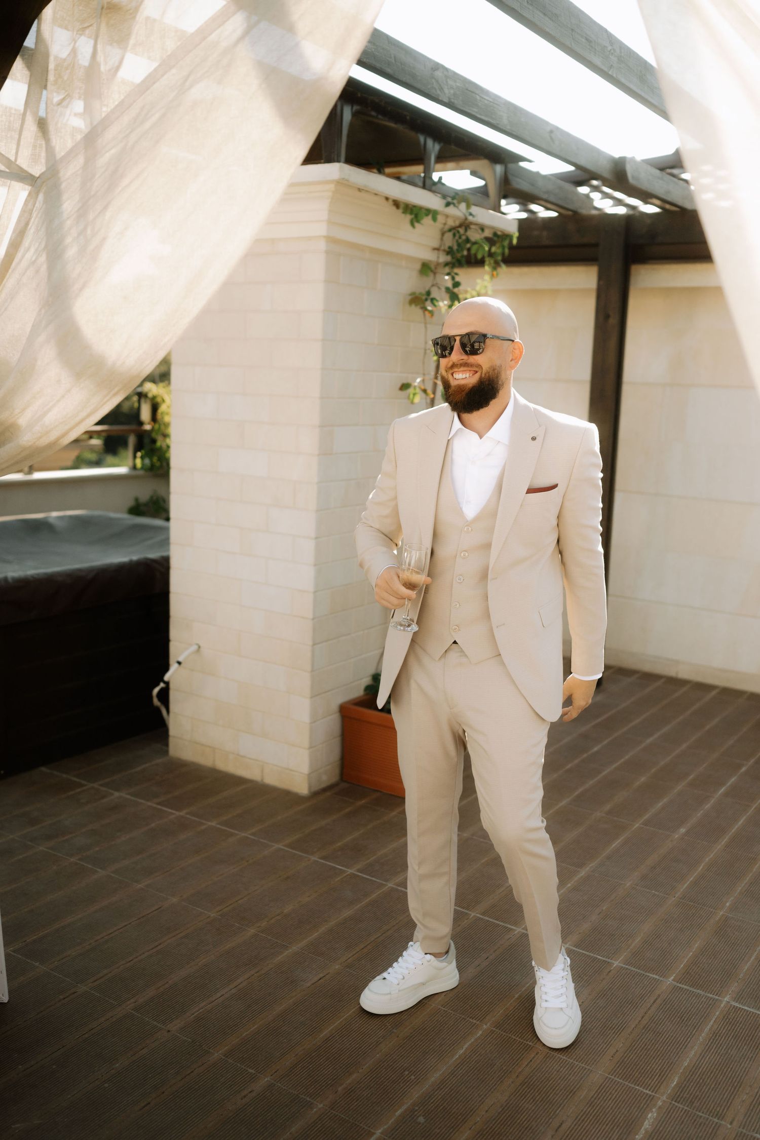 A person wearing a beige three-piece suit and white sneakers stands confidently on a tiled patio with flowing curtains.
