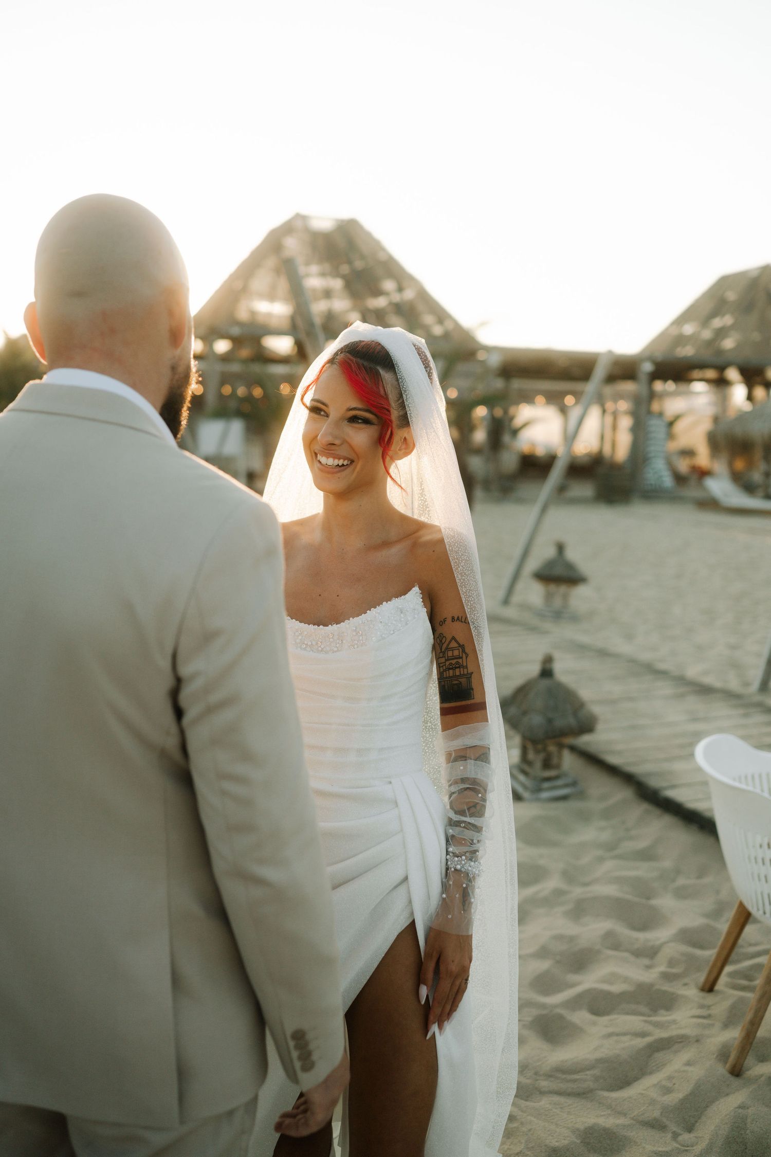 Newlyweds pose together on a sandy beach with thatched umbrellas and warm evening light.