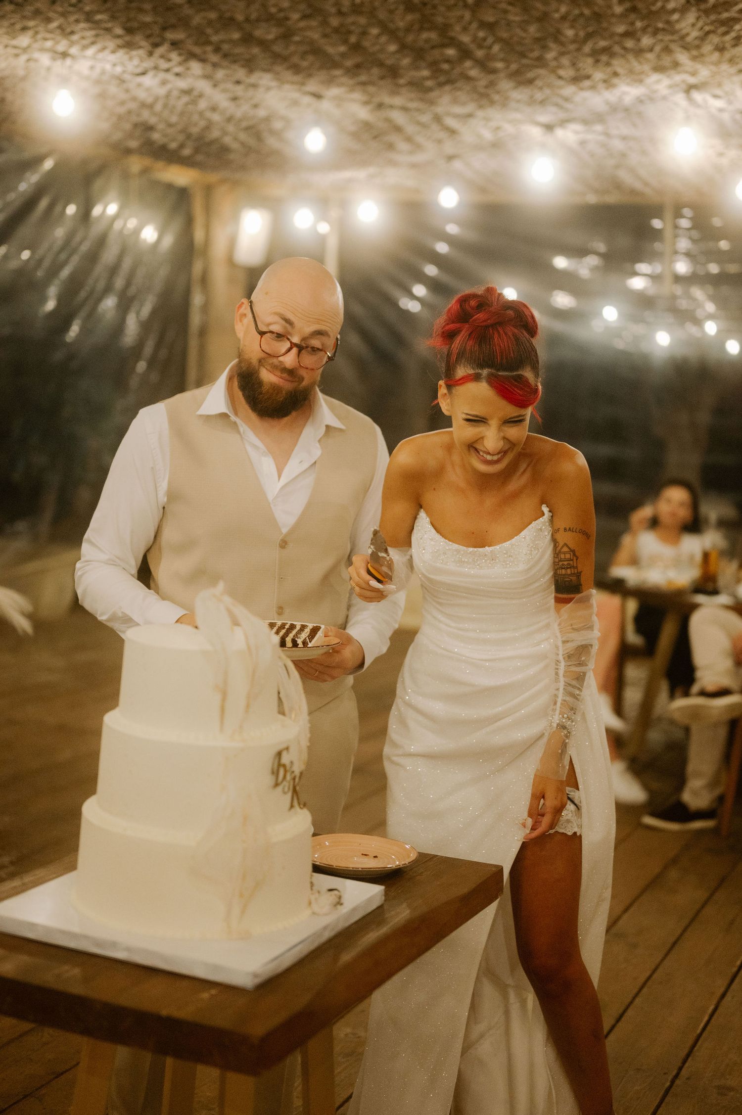 A couple cuts their elegant white wedding cake together under string lights at an outdoor evening reception.