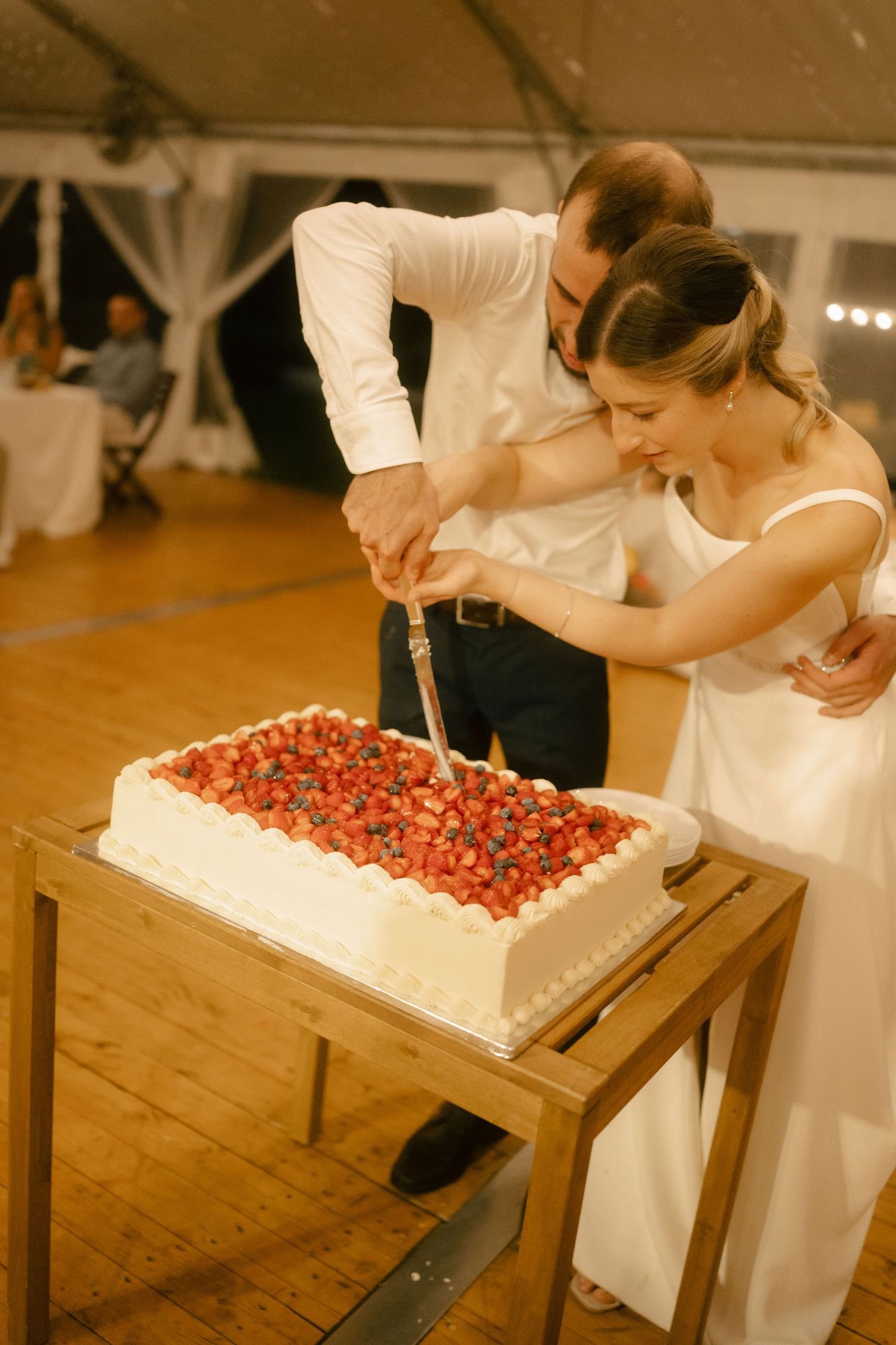 A couple in white attire cuts into a large white wedding cake decorated with fresh strawberries at their reception.
