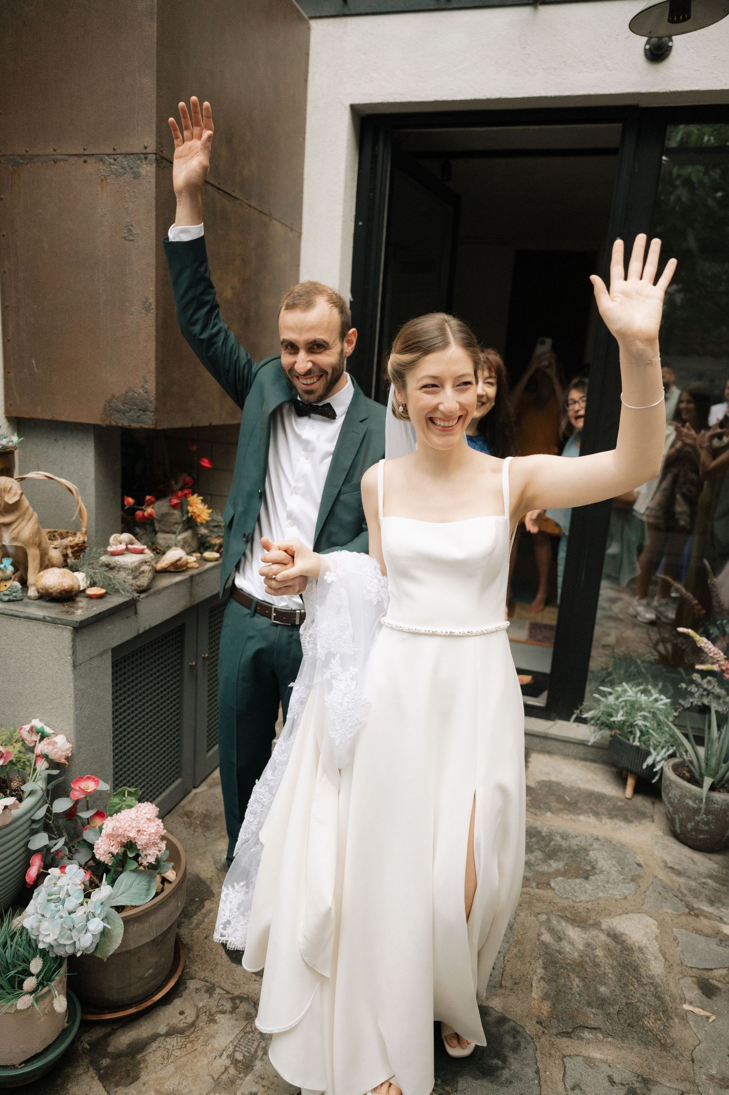 Wedding celebration with people in white and green attire raising their hands joyfully outside a building.