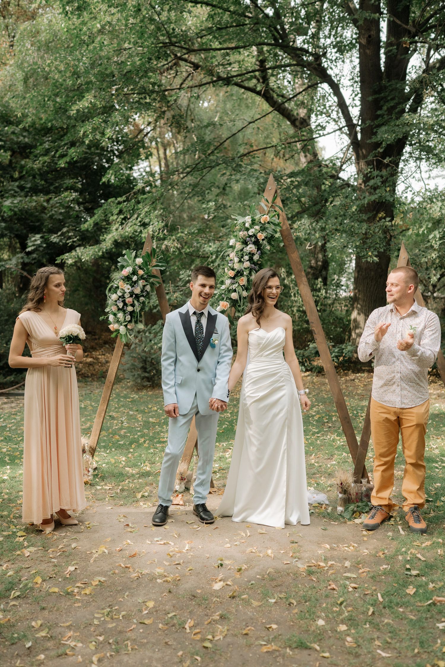 A playful wedding party poses in a natural outdoor setting with trees and scattered leaves on the ground.