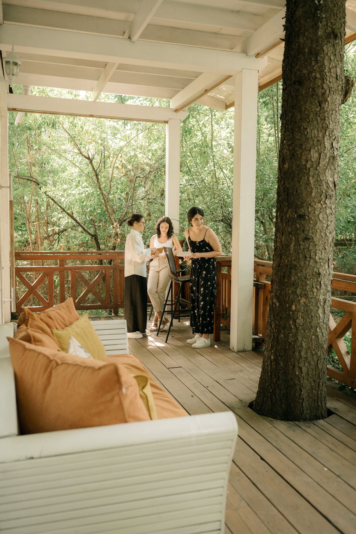 A group gathers on a rustic wooden deck porch with white pillars surrounded by lush greenery and comfortable seating.