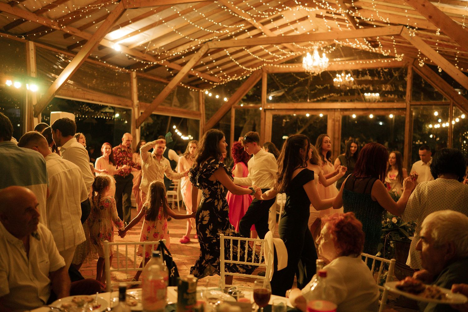Wedding guests dance and celebrate on an illuminated dance floor under a thatched roof pavilion at night.