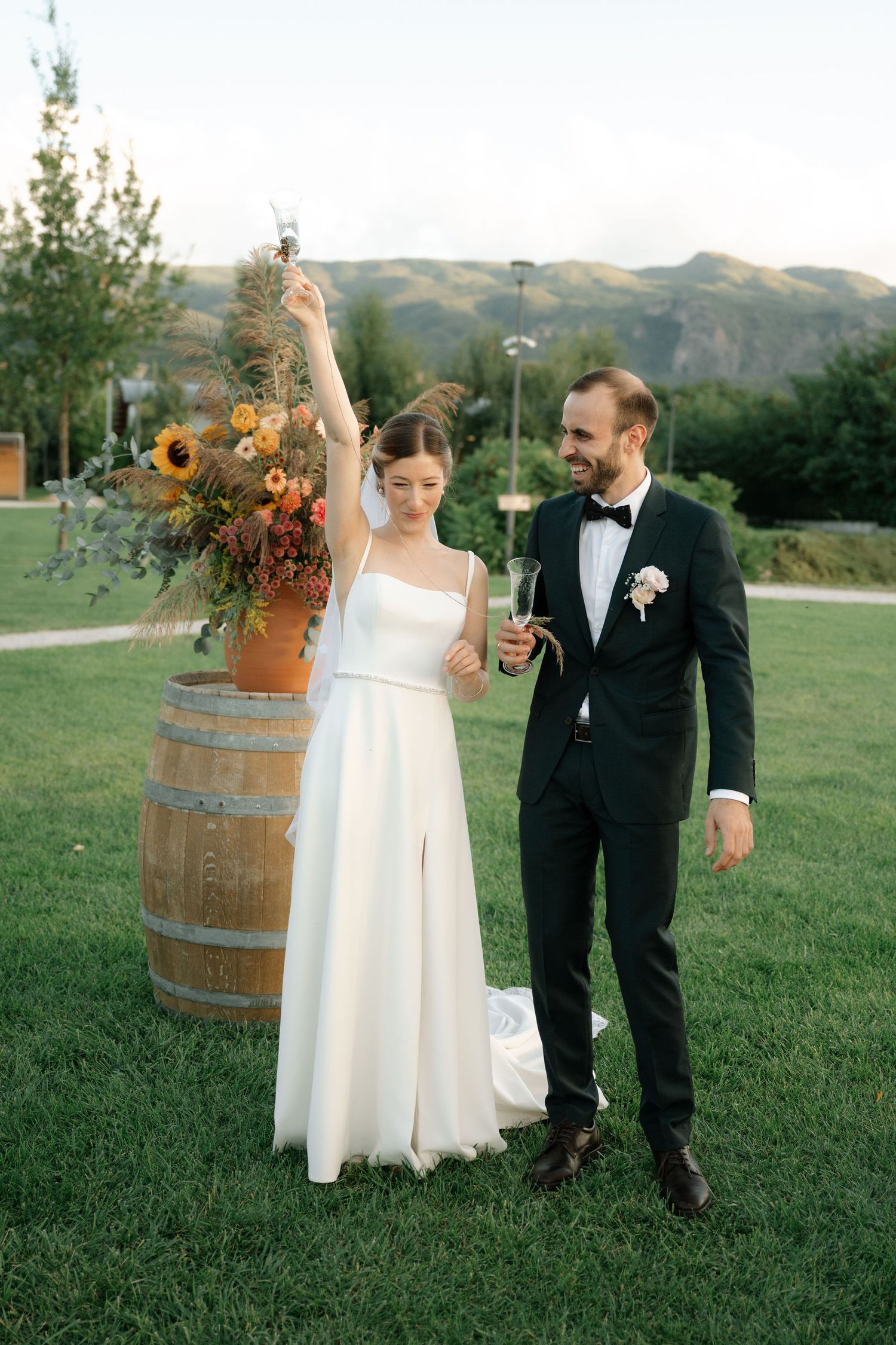 A couple in wedding attire poses together on a grassy hillside with rustic wine barrels and sunflowers at sunset.