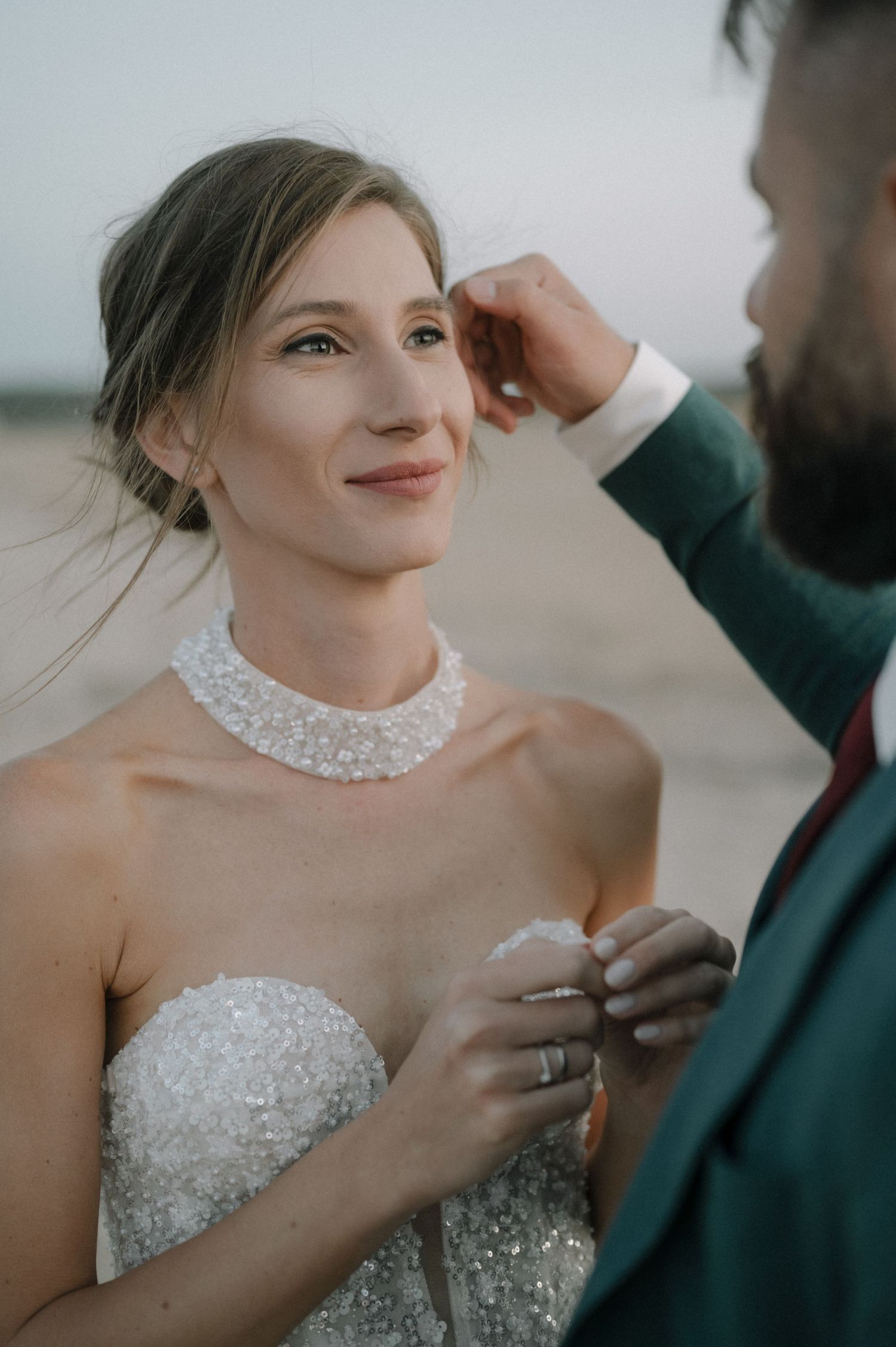 Groom tenderly brushes brides hair as she smiles wearing beaded gown and pearl necklace on beach.