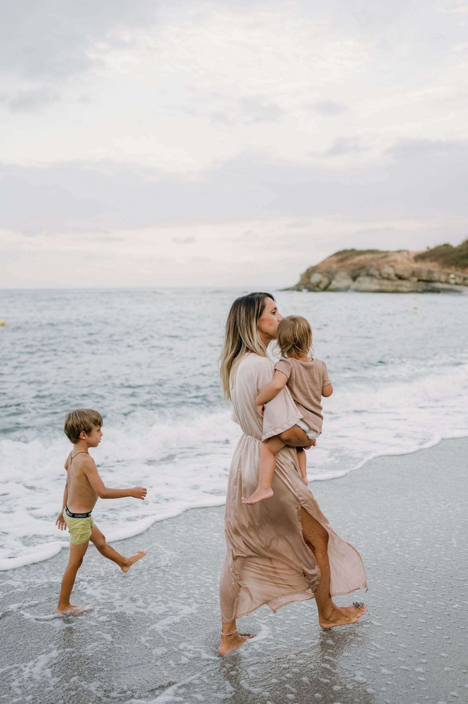 A mother in a flowing pink dress kisses her toddler while walking barefoot on a beach with her young son.