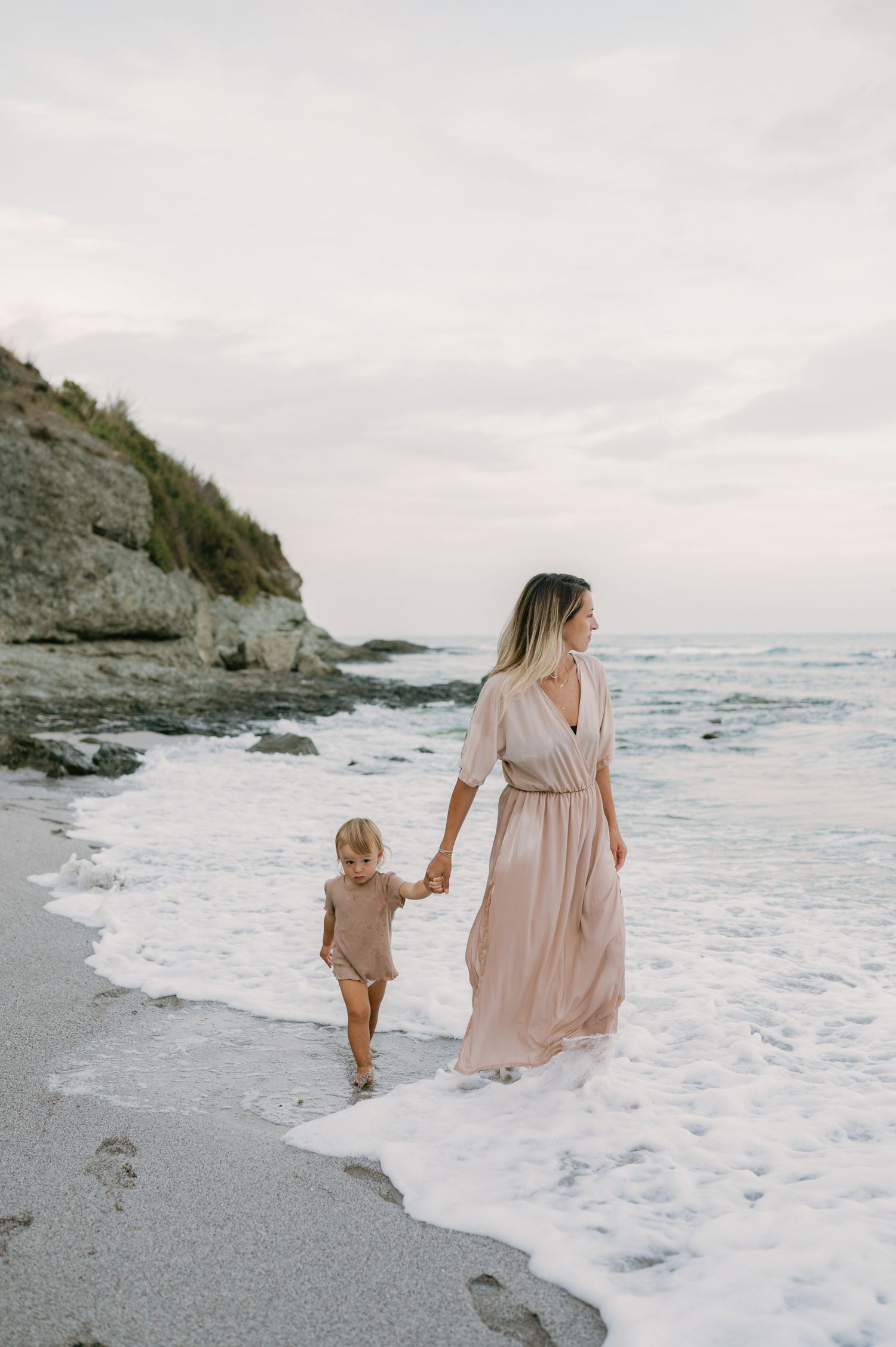 A mother in a flowing pink dress holds hands with her toddler while walking along a rocky beach shoreline.