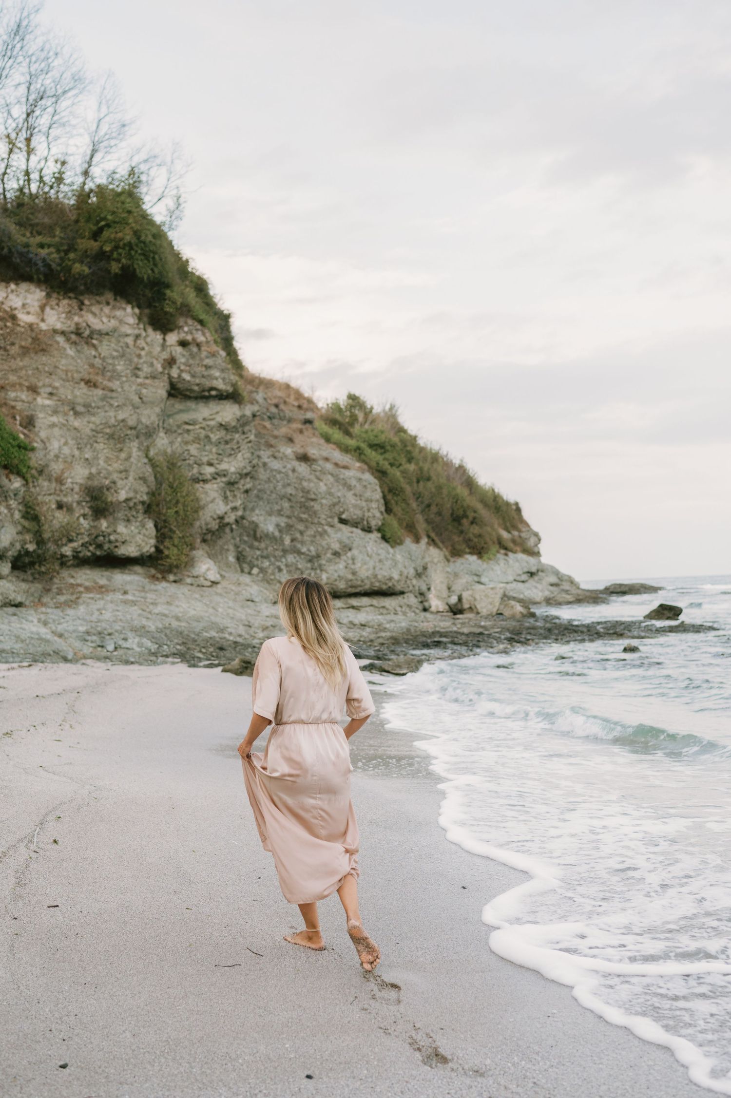 Woman in pink maxi dress walking barefoot on sandy beach with rocky cliffs and ocean waves.
