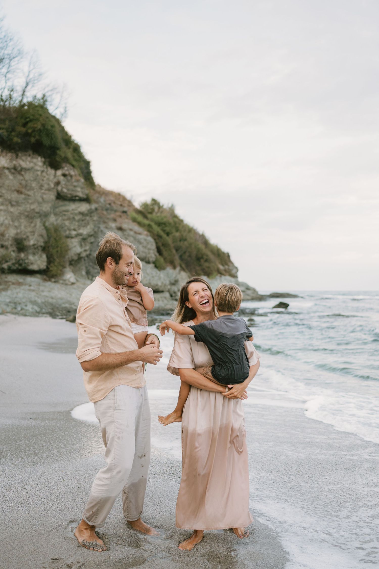 Happy family of four laughing and playing on a sandy beach at dusk, parents holding young children near ocean waves.