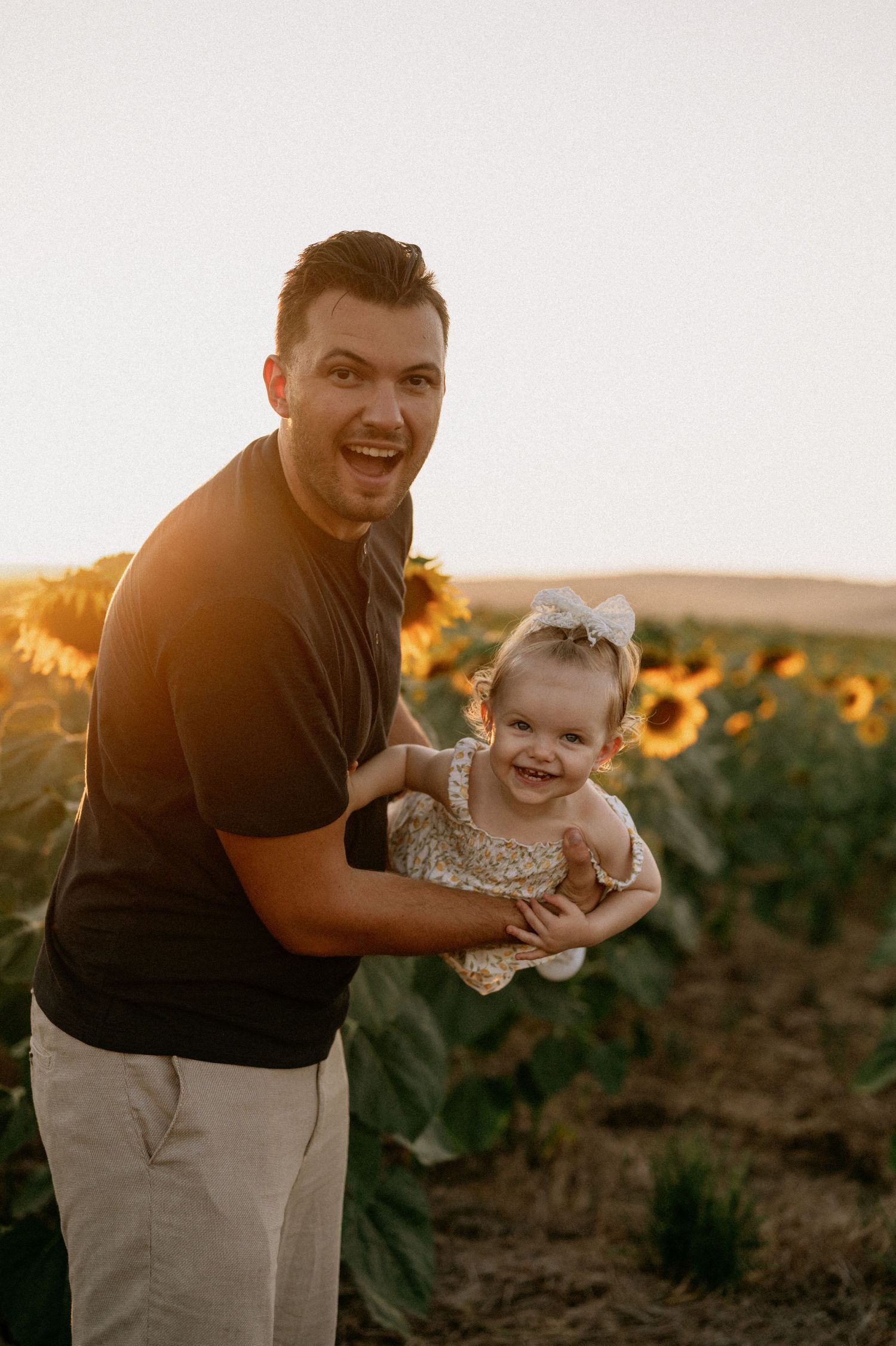 A joyful man holds a smiling baby girl in a sunflower field during golden hour sunset.
