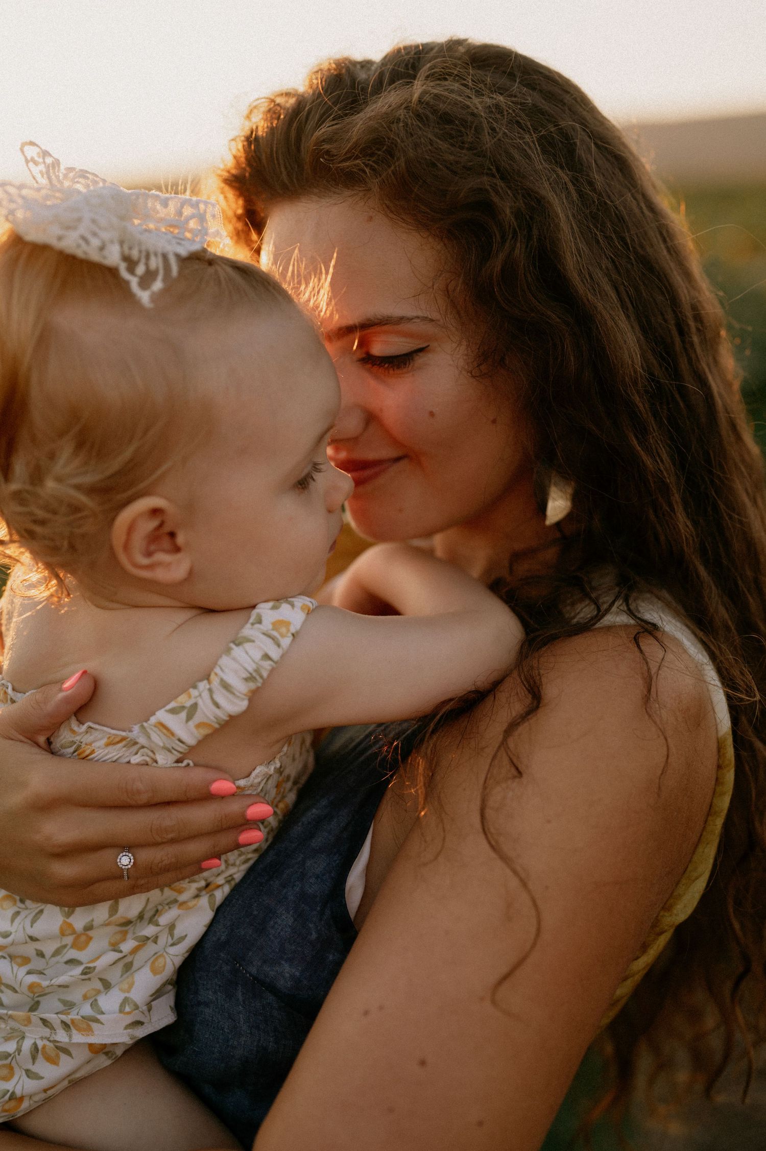 A young mother with curly hair tenderly holds her baby girl in a golden sunset field.