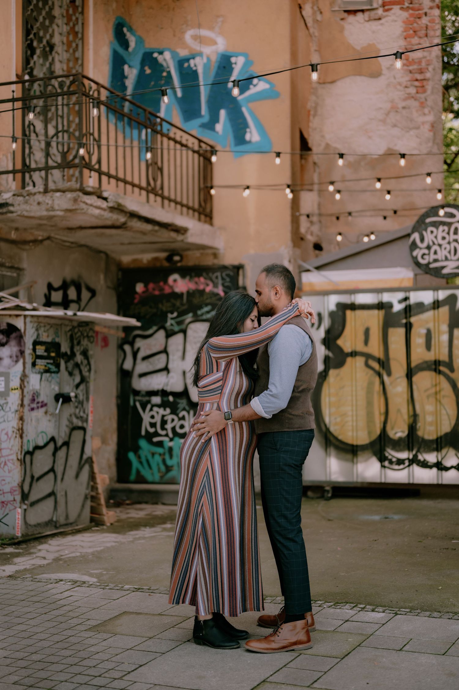 Couple embracing romantically in front of colorful graffiti wall with string lights in urban setting.