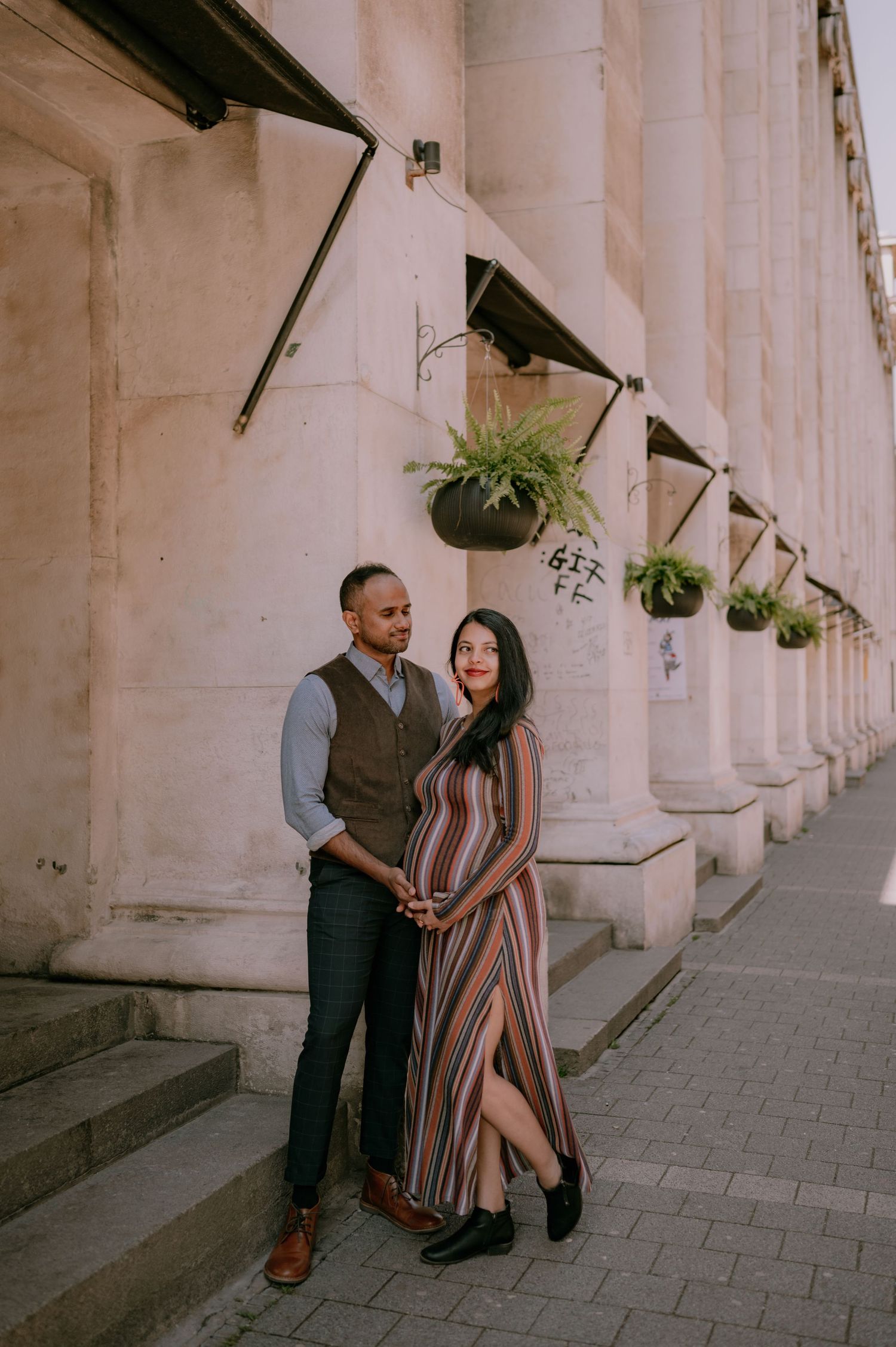 Couple posing together outside a historic stone building with hanging fern planters, woman in striped maxi dress showing baby bump.