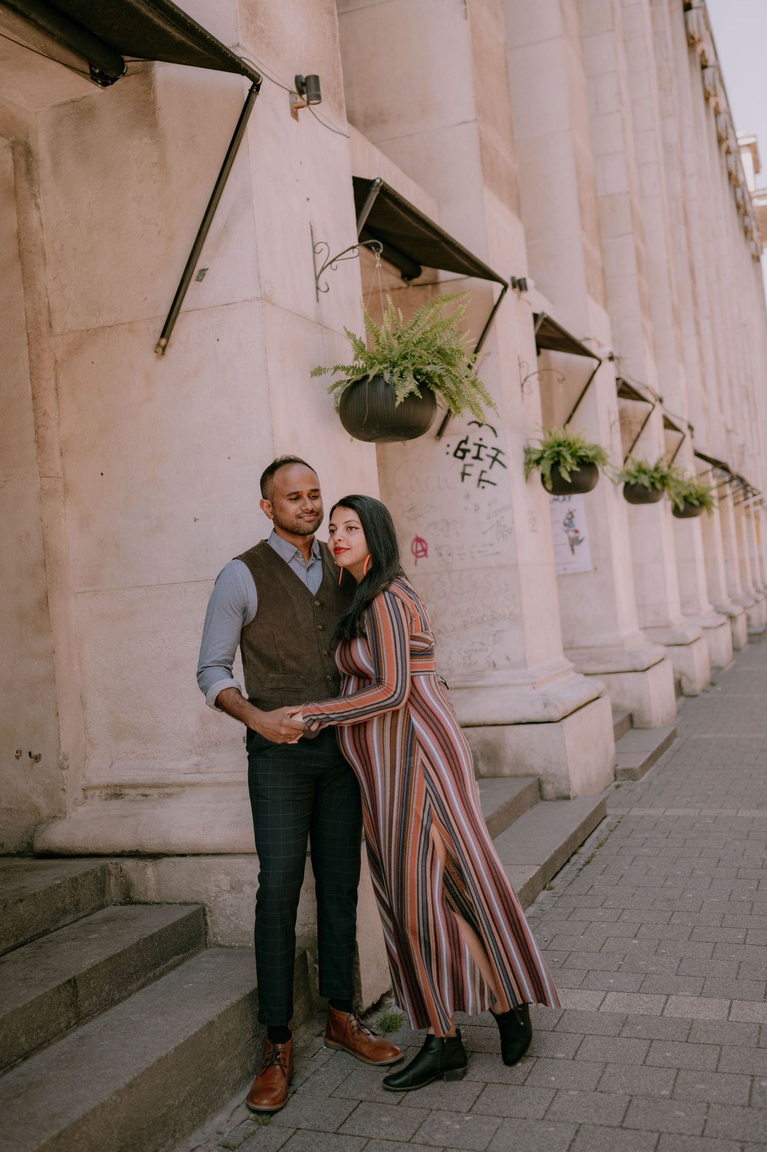 Couple posing together against a stone building with hanging fern planters on a city street.
