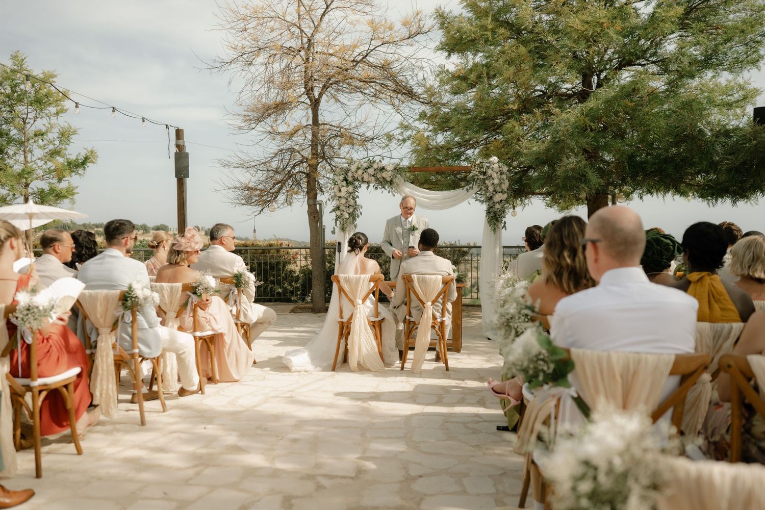 Outdoor wedding ceremony with guests seated on wooden chairs under string lights and trees on a sunny day.
