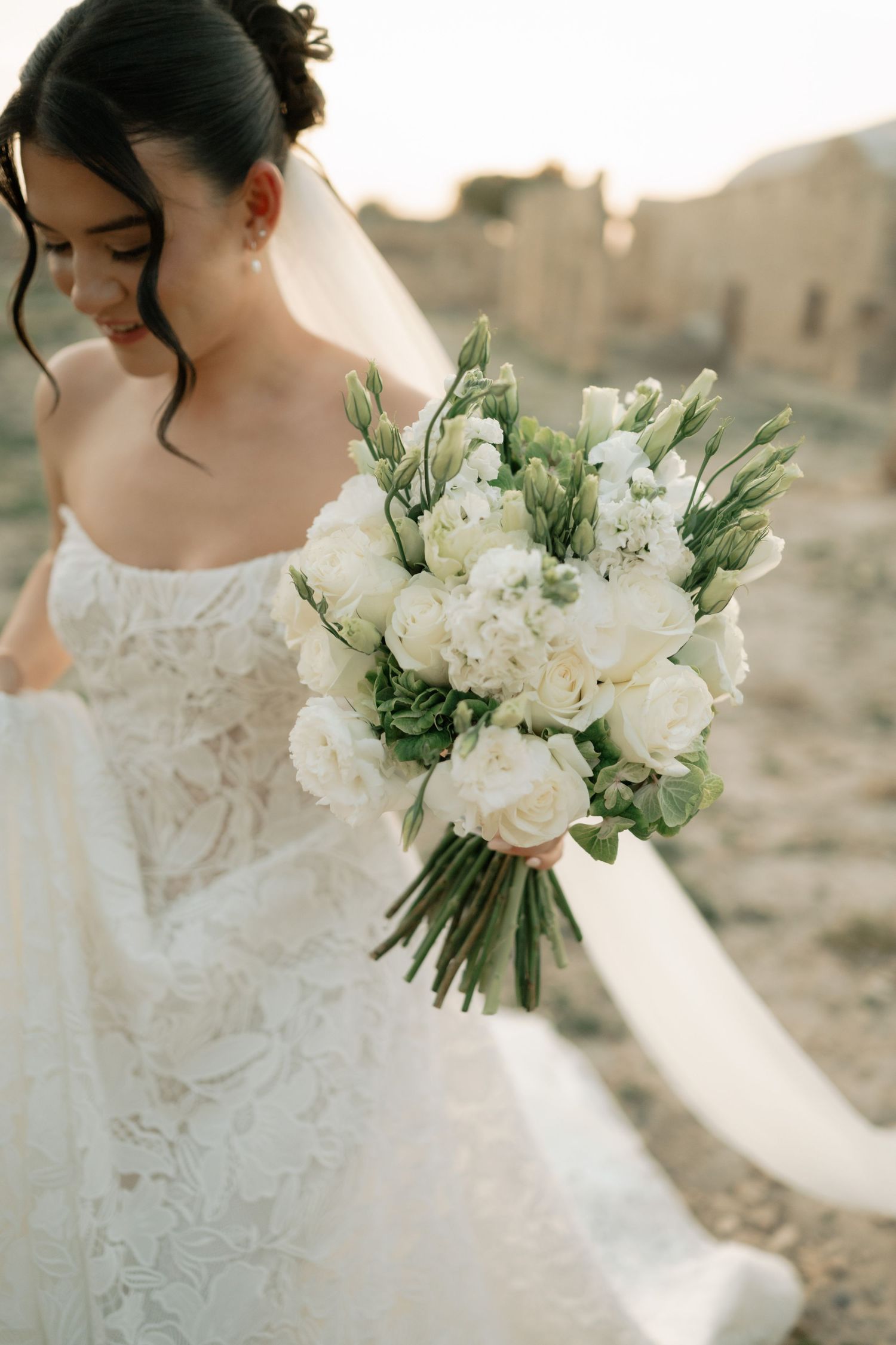 Bride in lace wedding dress holds elegant white rose bouquet with greenery outdoors in golden light.