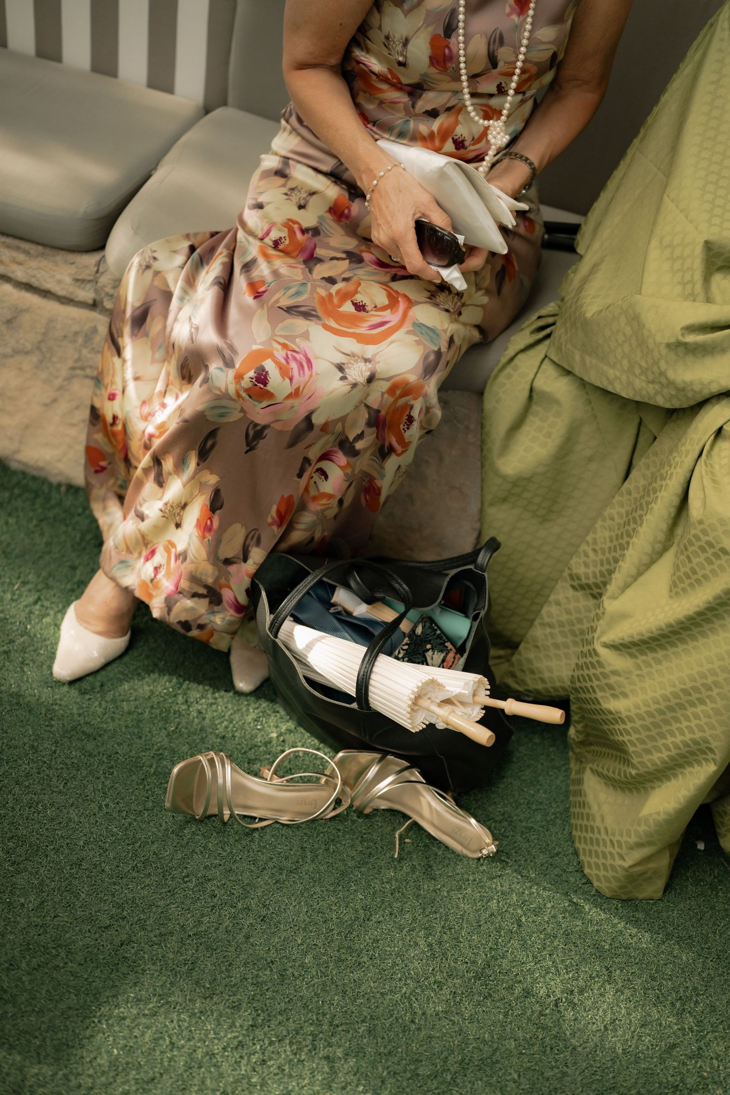 Woman in floral dress seated with open handbag, white fan, and strappy heels on green carpet floor.