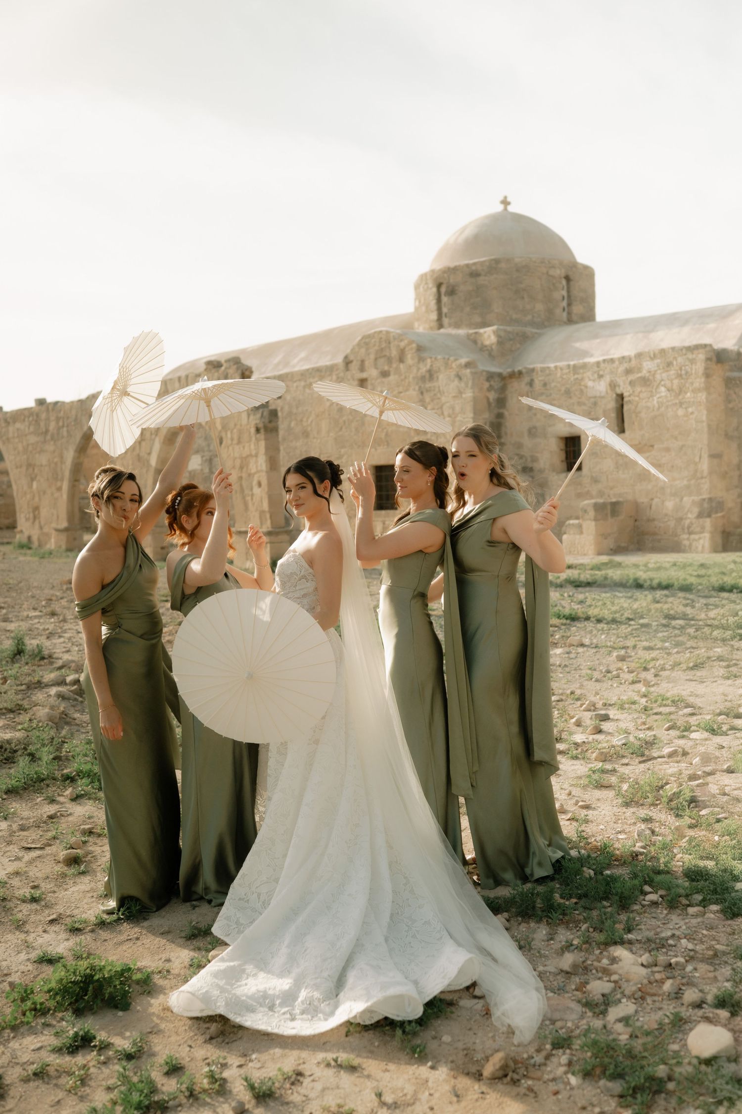 Bride and bridesmaids in sage green gowns holding white parasols near ancient stone ruins on a sunny day.