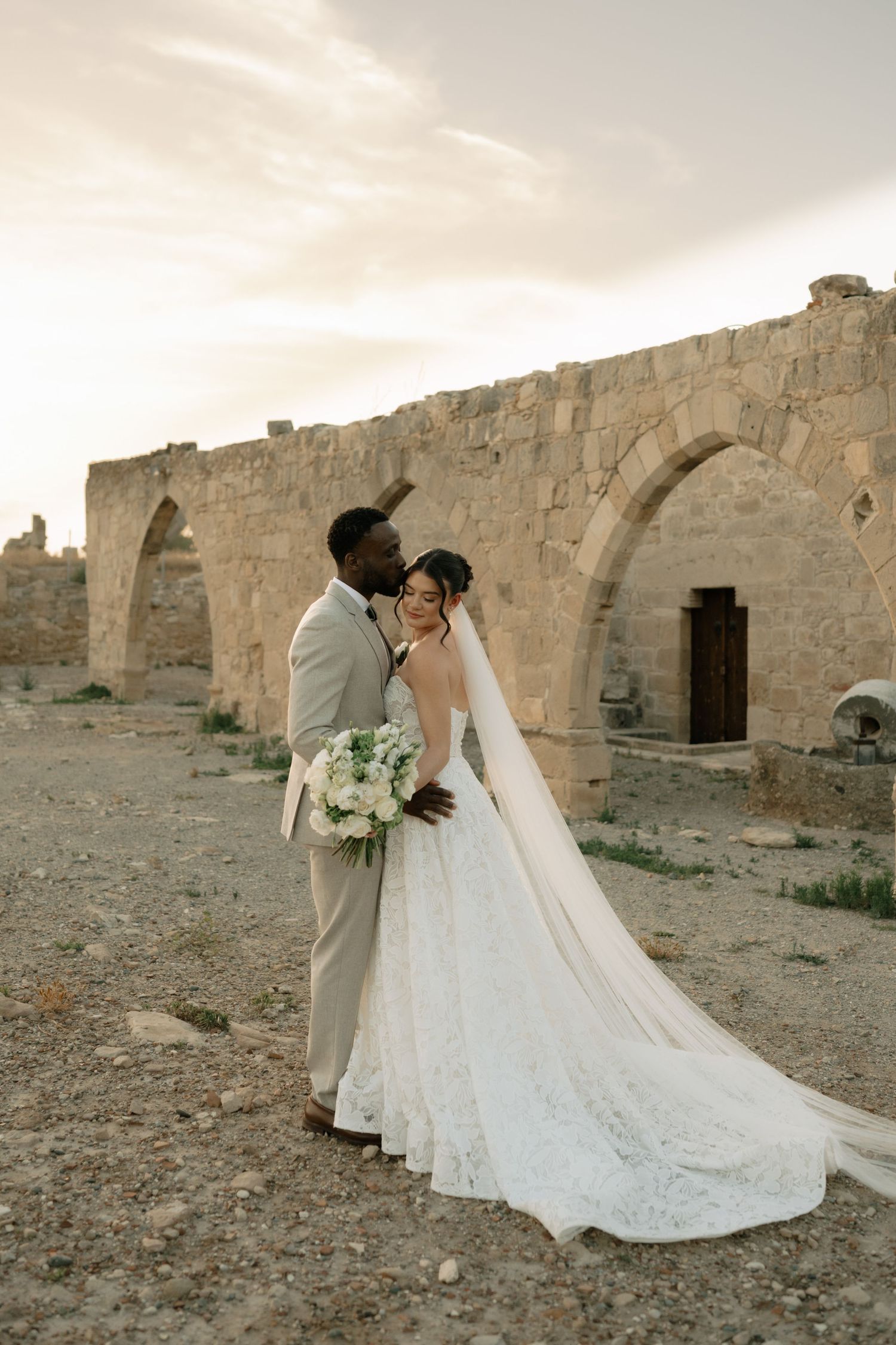 Interracial couple in wedding attire posing before ancient stone arches at sunset, bride holding white bouquet.