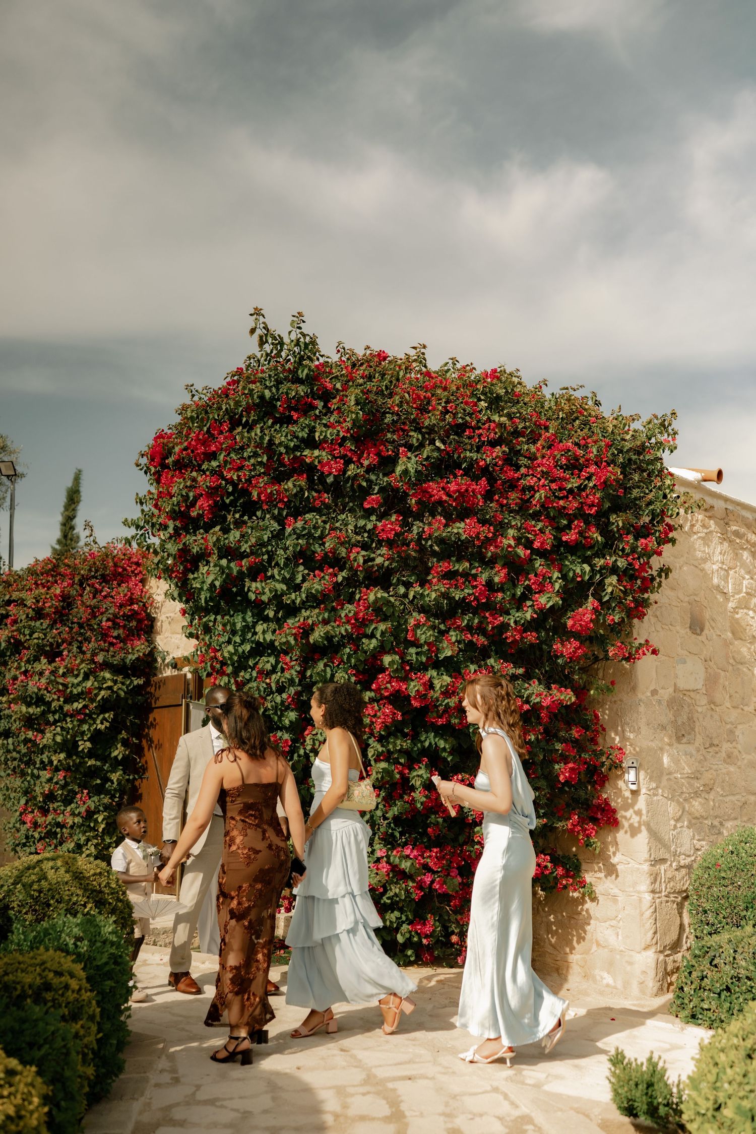 Women in elegant dresses walk past a stunning red bougainvillea wall at a Mediterranean stone venue.