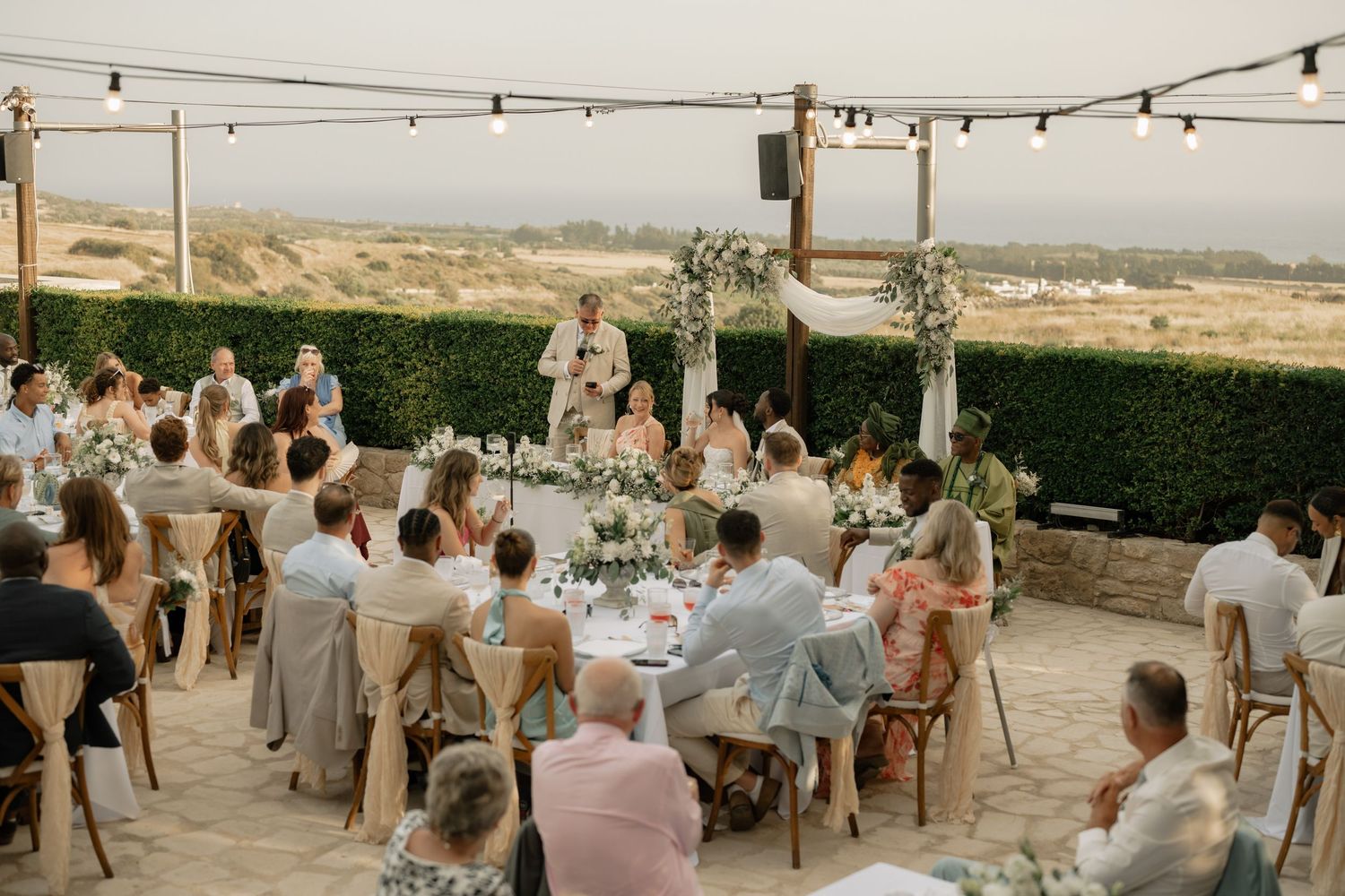 Outdoor wedding reception with guests seated at decorated tables under string lights, with a scenic desert landscape backdrop.