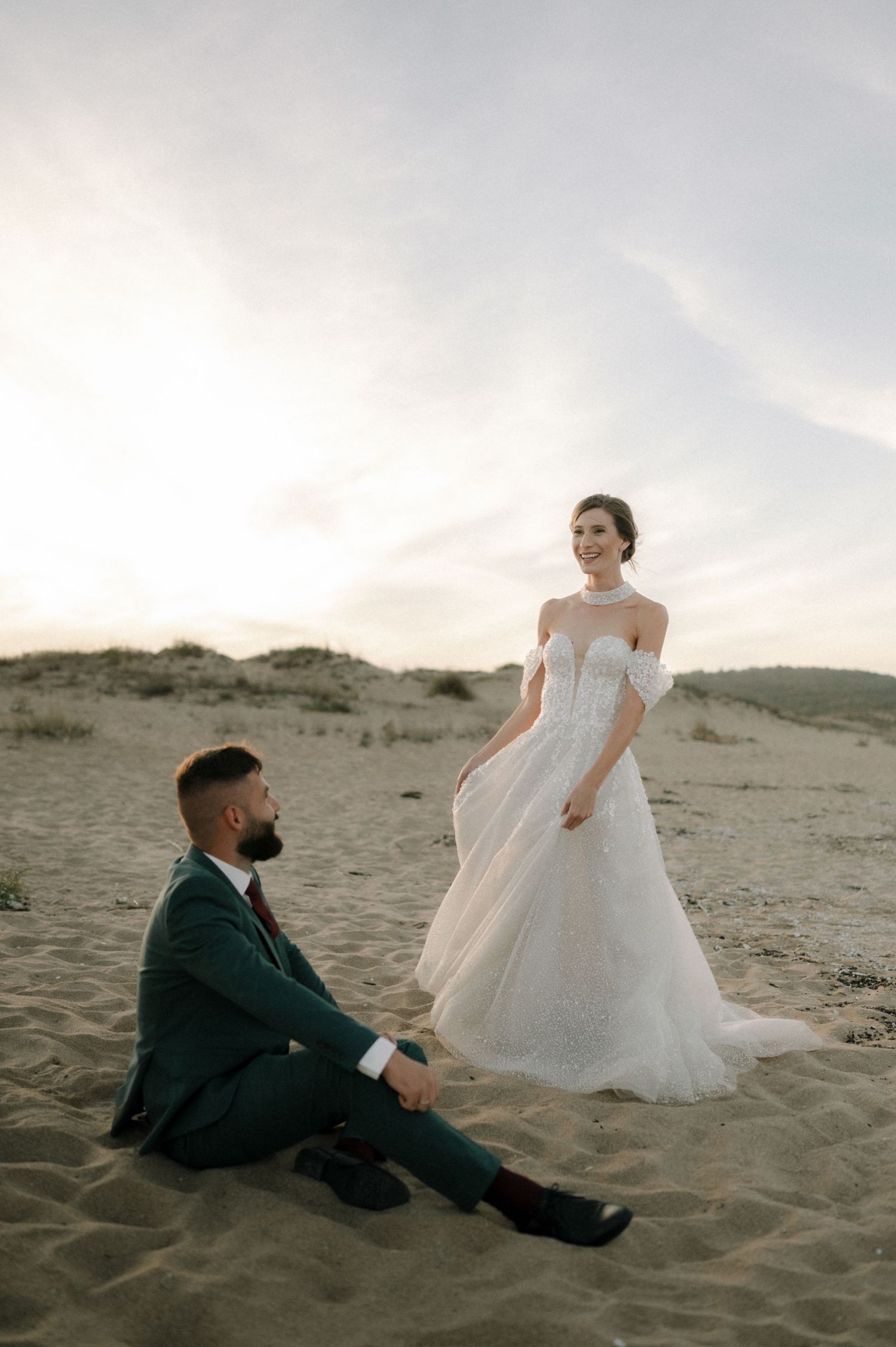 Bride in white off-shoulder gown stands on sandy dunes as groom in green suit sits looking up at her at sunset.