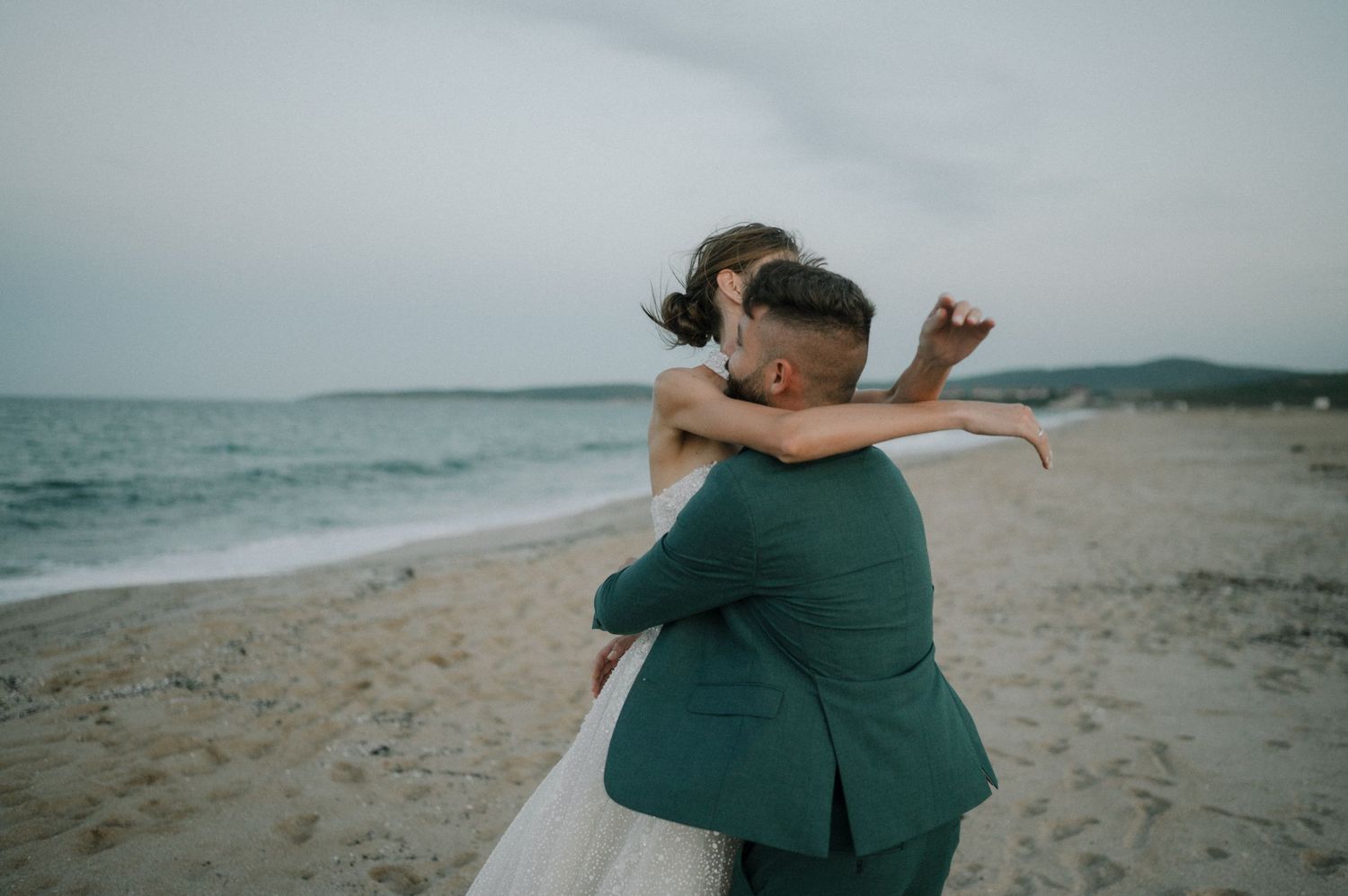 Bride and groom embracing on a sandy beach at dusk, groom in green suit, romantic wedding photo.