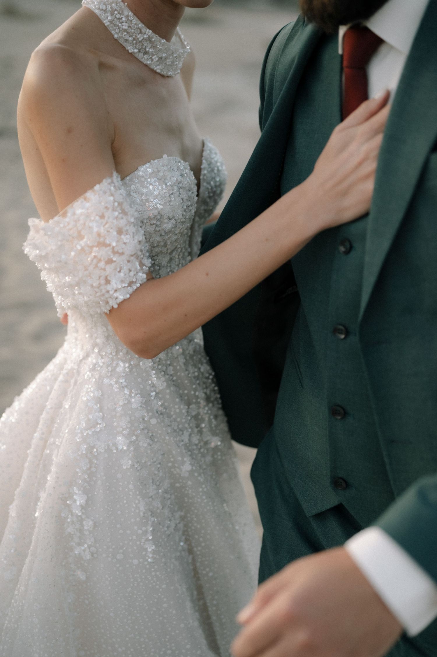 Bride in off-shoulder beaded gown stands beside groom in dark green suit with red tie at wedding.