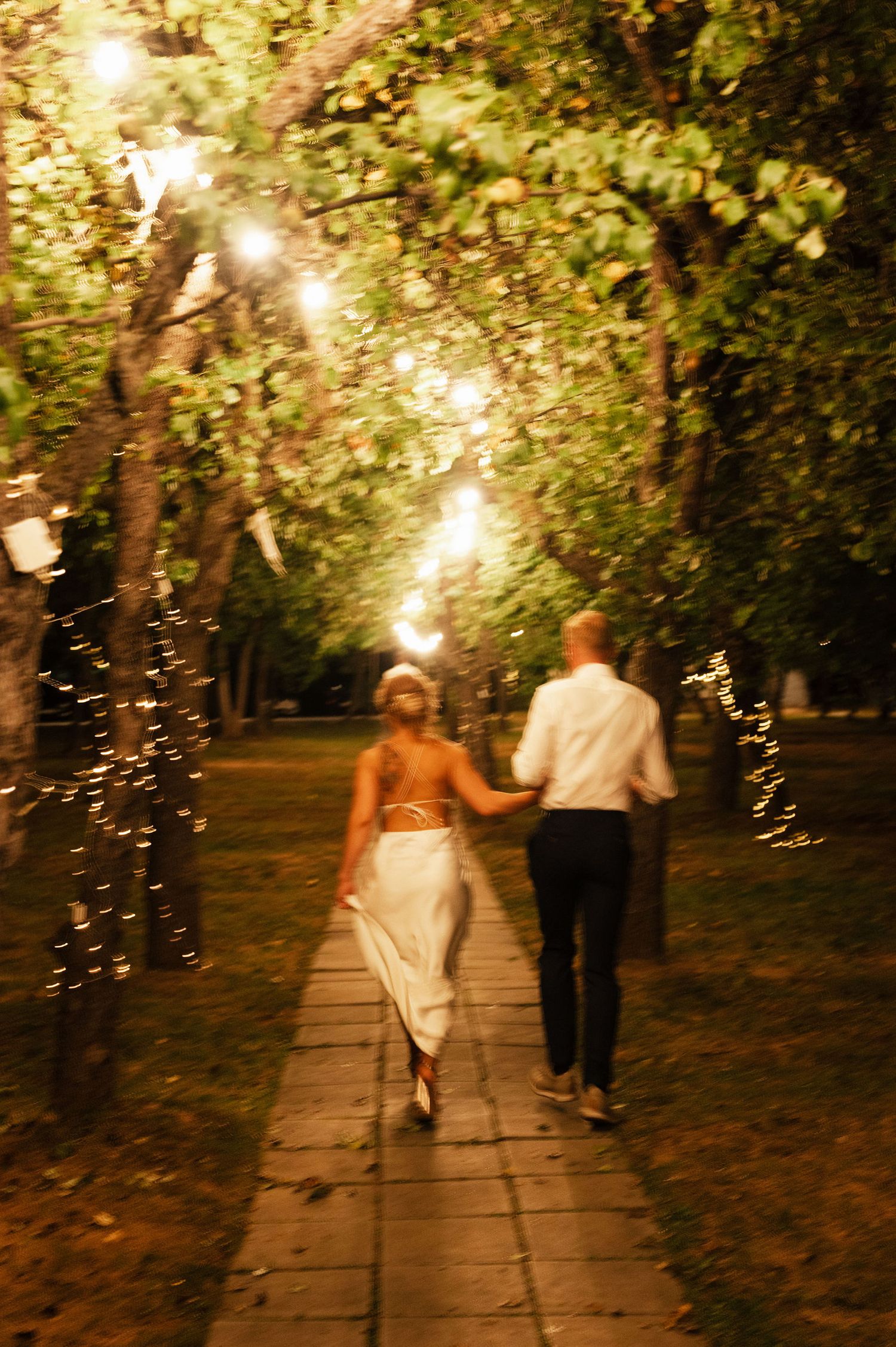 A couple walks along an illuminated garden path at dusk with string lights glowing in the trees above.