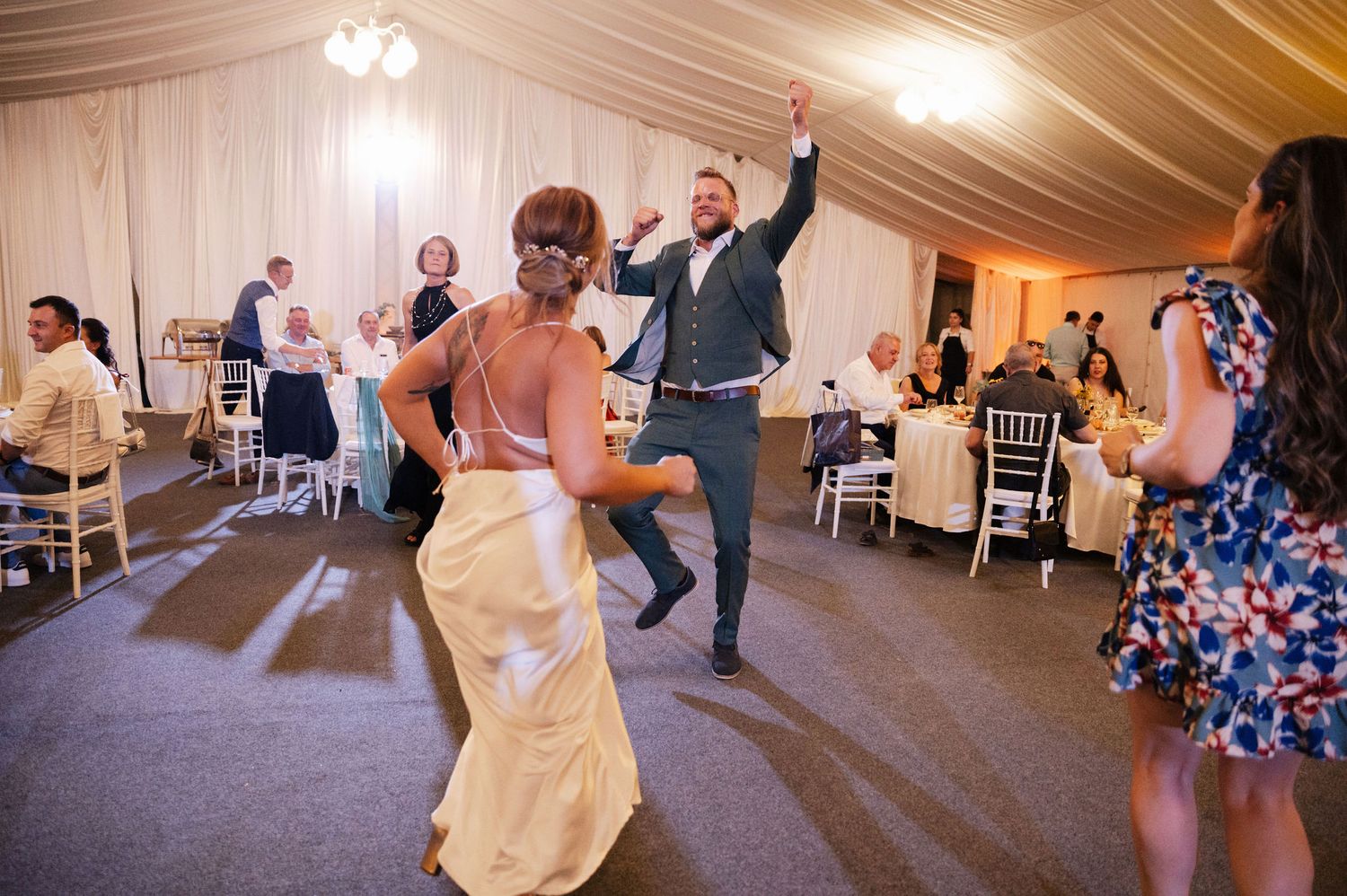 Wedding guests dance together on a carpeted floor under a white tent during an evening reception celebration.