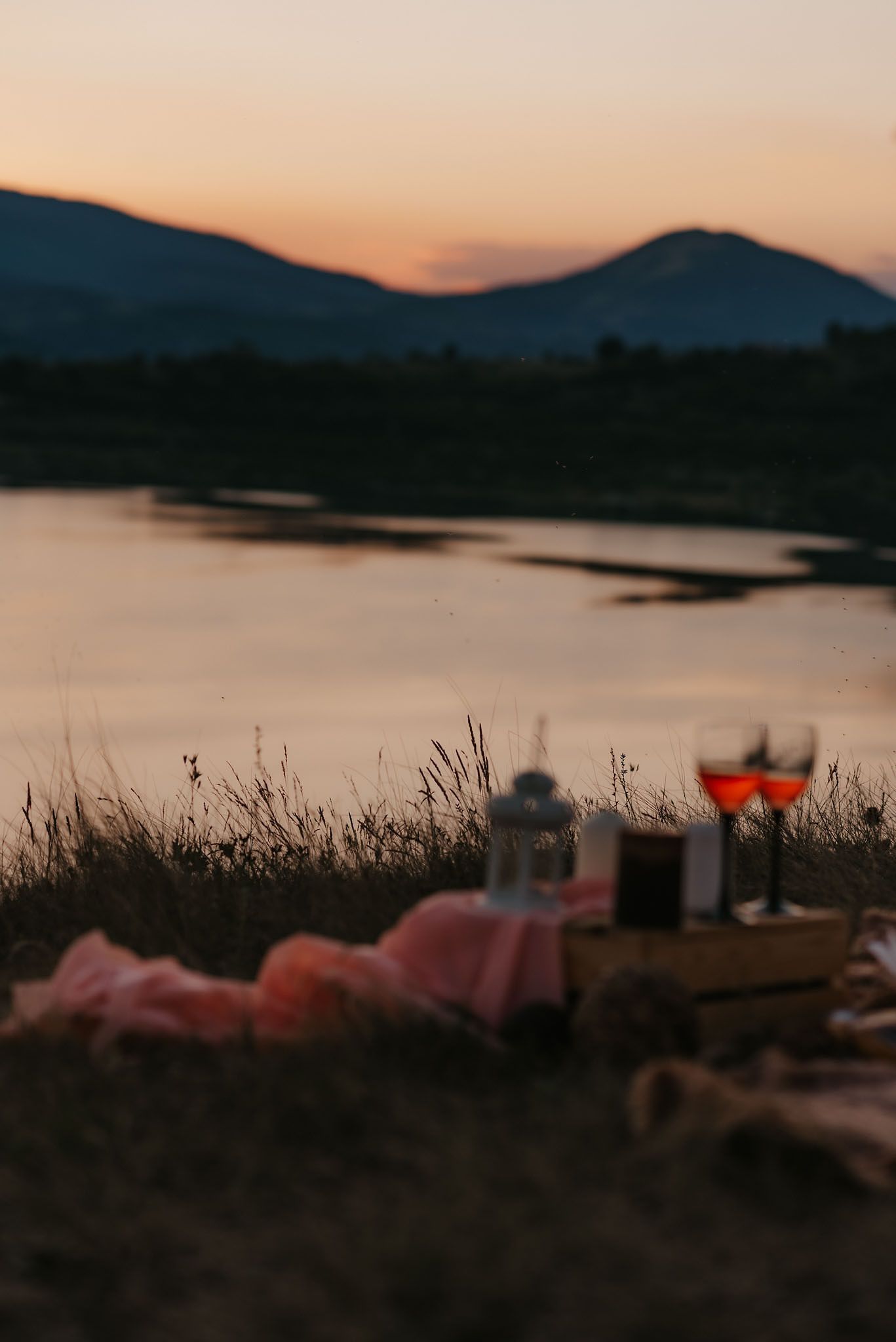 A romantic lakeside picnic setup at sunset with blankets and decorations along the shoreline.