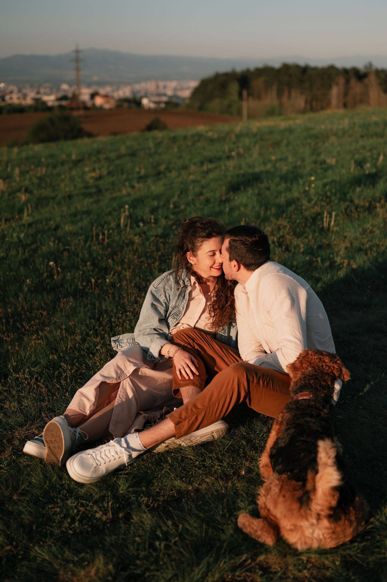 A couple in white clothing lying together on a grassy hillside at sunset with a city view in the background.