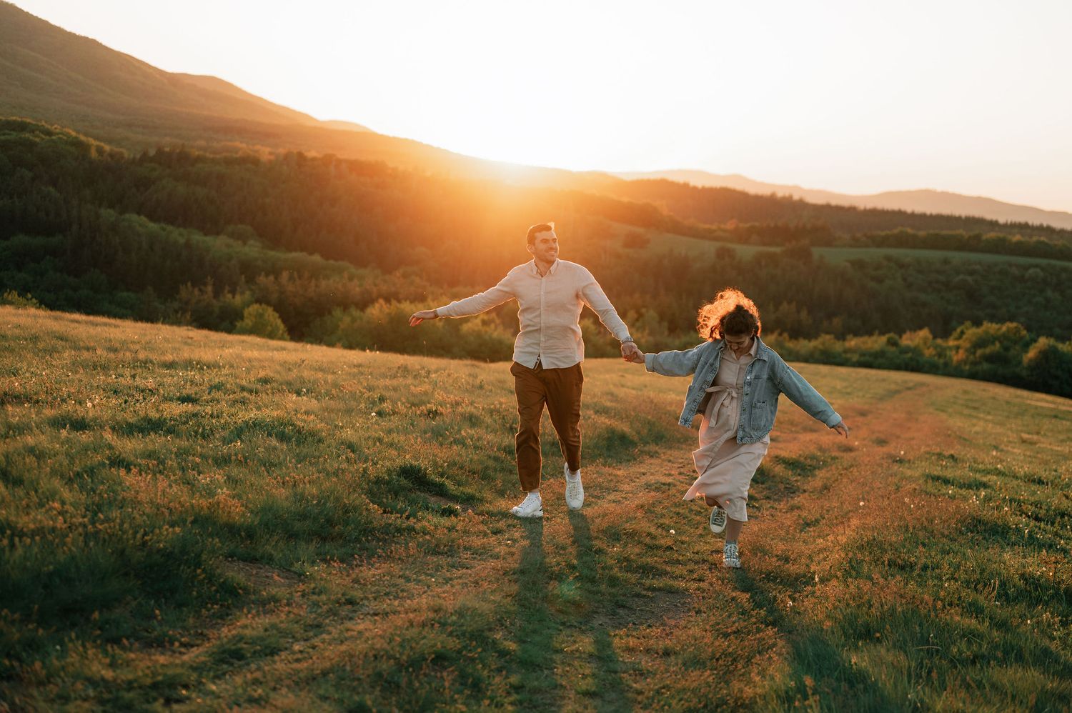 Two people dancing together in a grassy meadow at sunset with golden light illuminating the mountainous landscape.