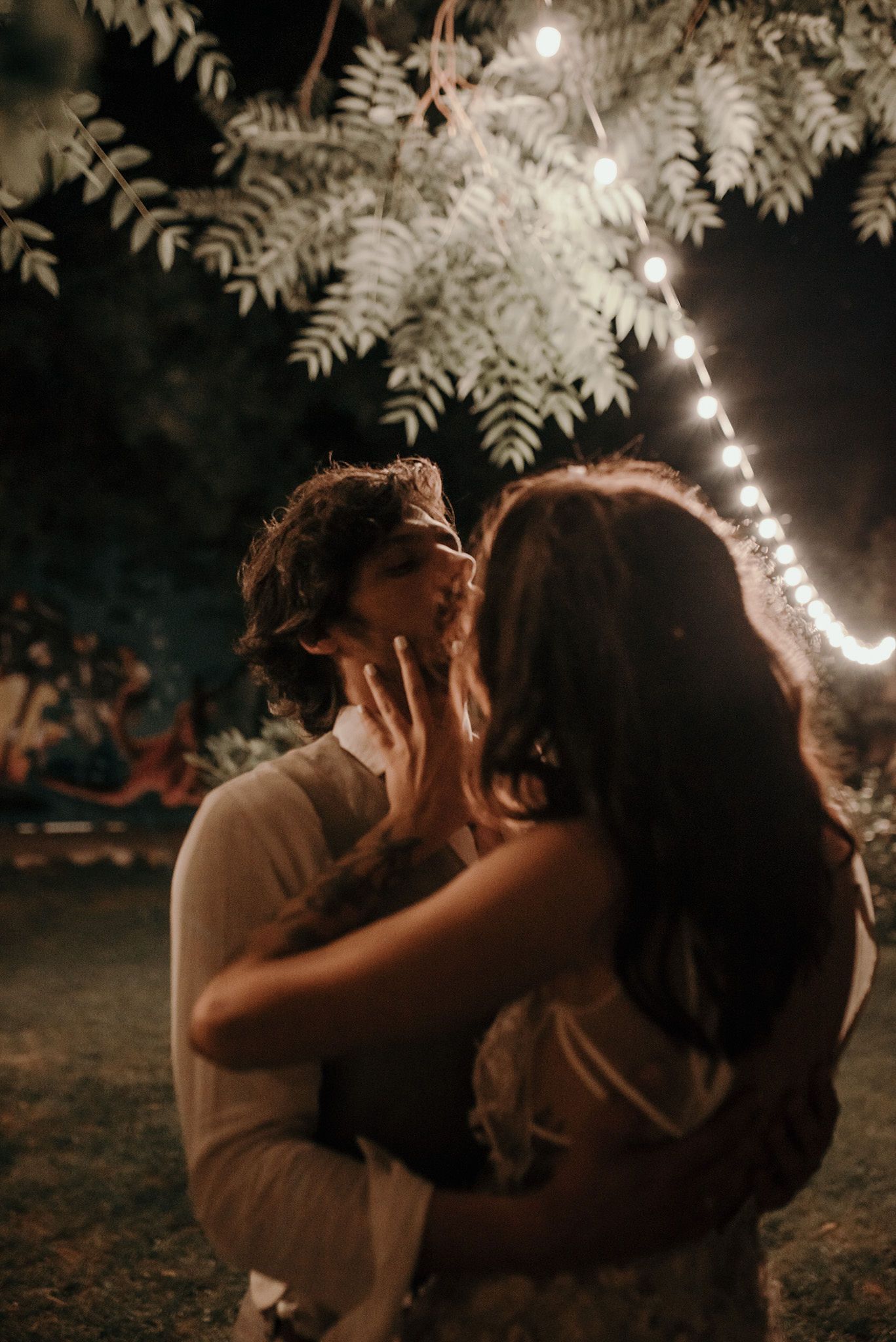 A romantic moment captured under string lights and leaves at an outdoor evening celebration.