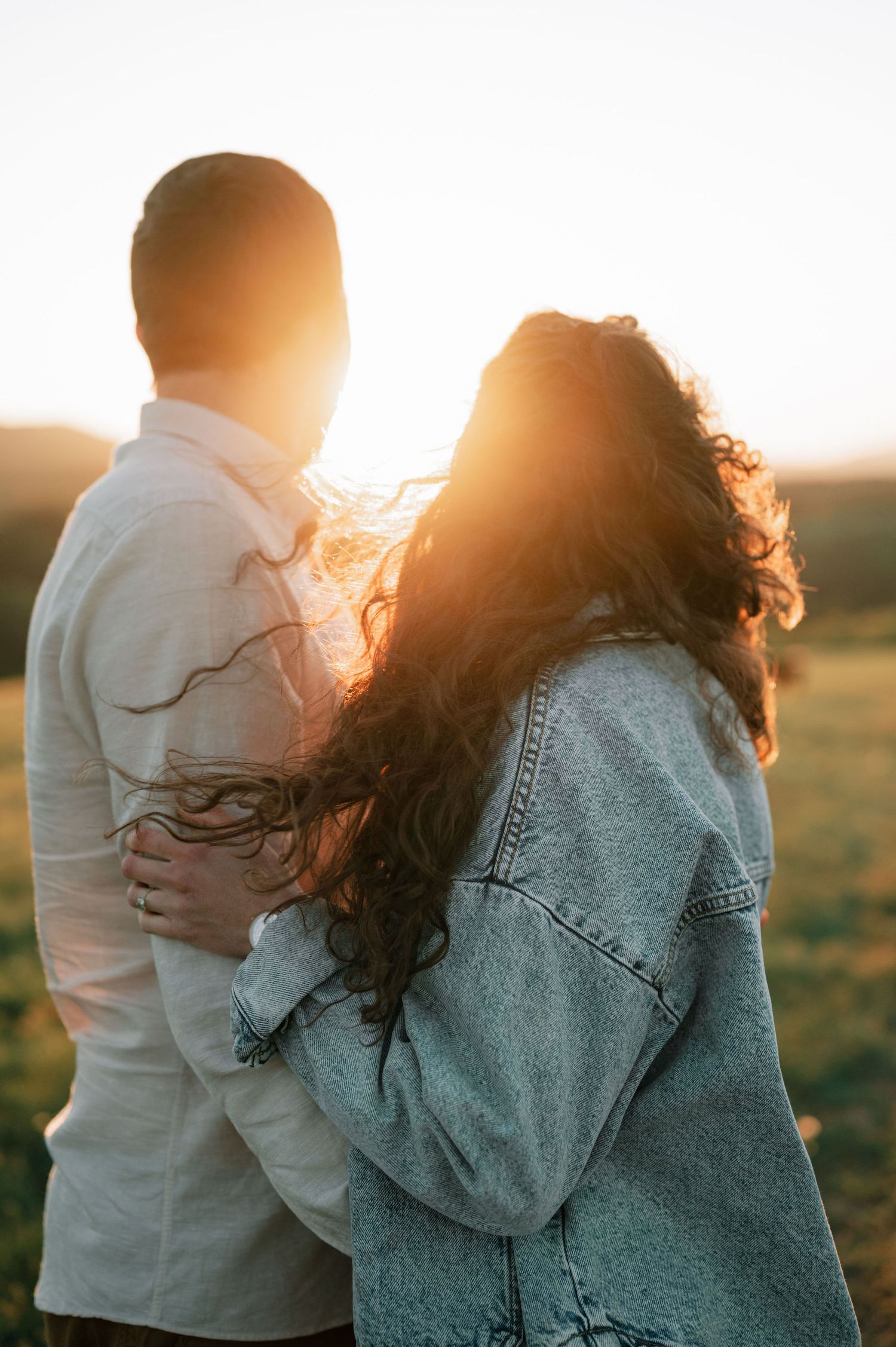 Couple embracing at sunset in a field wearing denim and white clothing as golden light glows behind them.