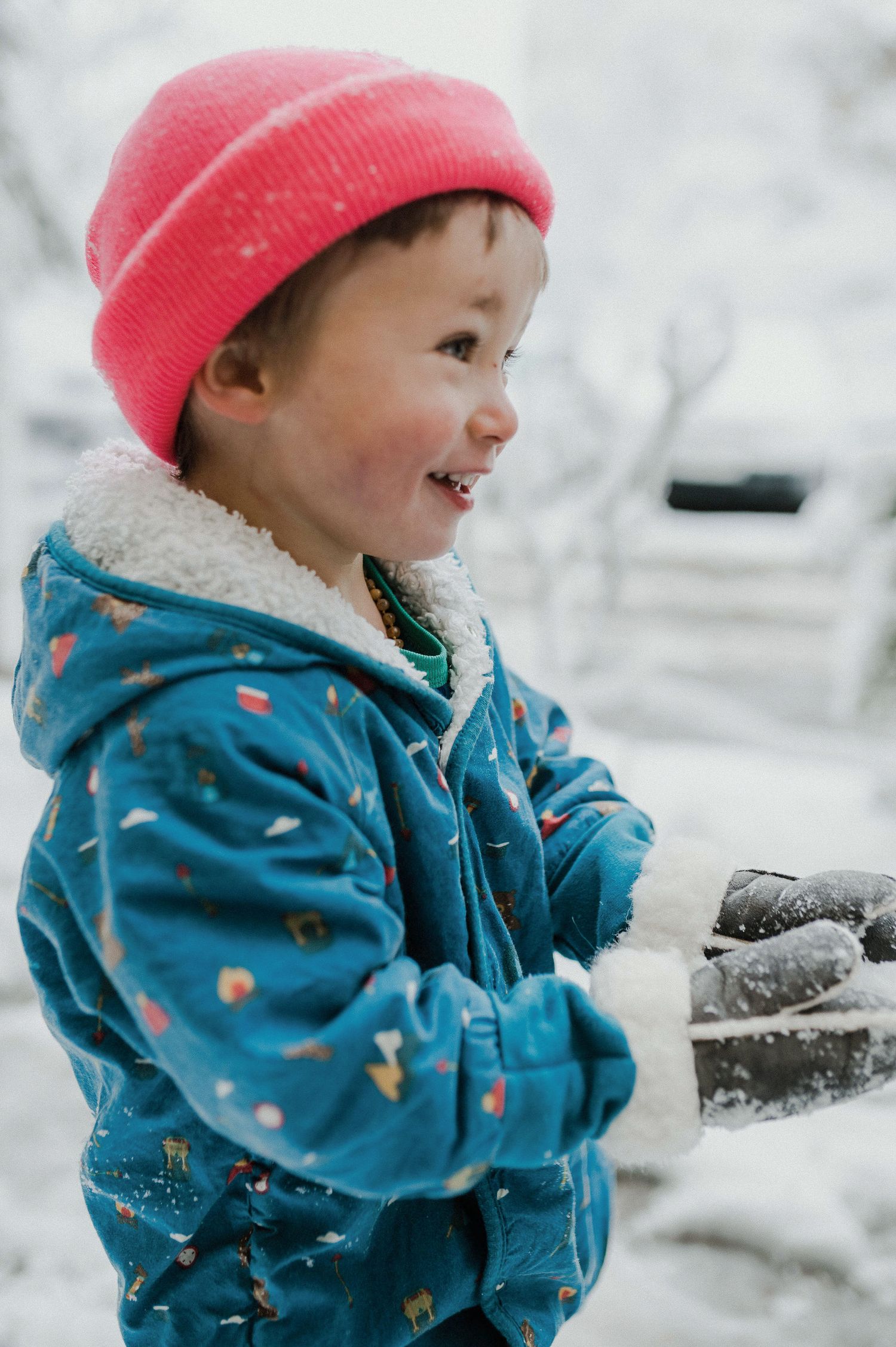 A small child in a teal winter coat and pink beanie plays joyfully in the snow on a winter day.