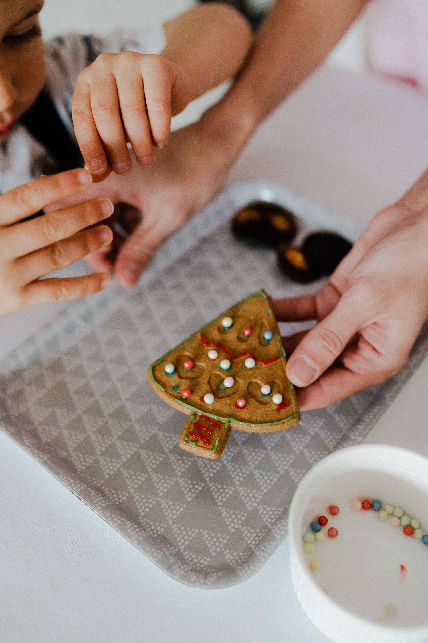 Hands decorating Christmas gingerbread cookies with colorful candy pieces on a patterned surface.
