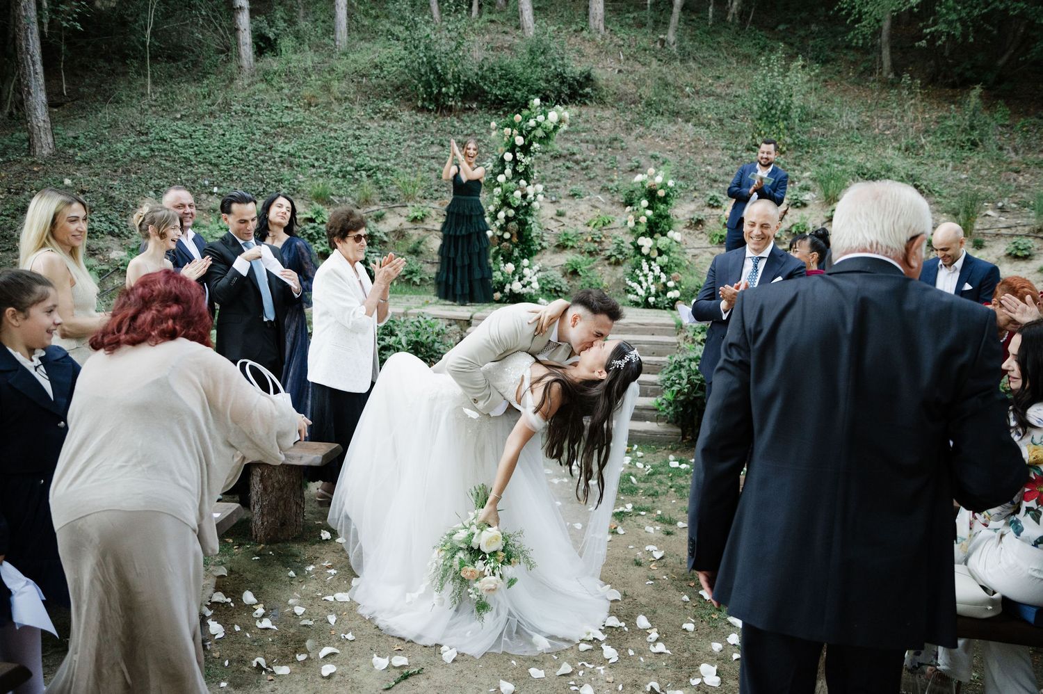 Wedding guests mingle during an outdoor ceremony in a scenic woodland setting.