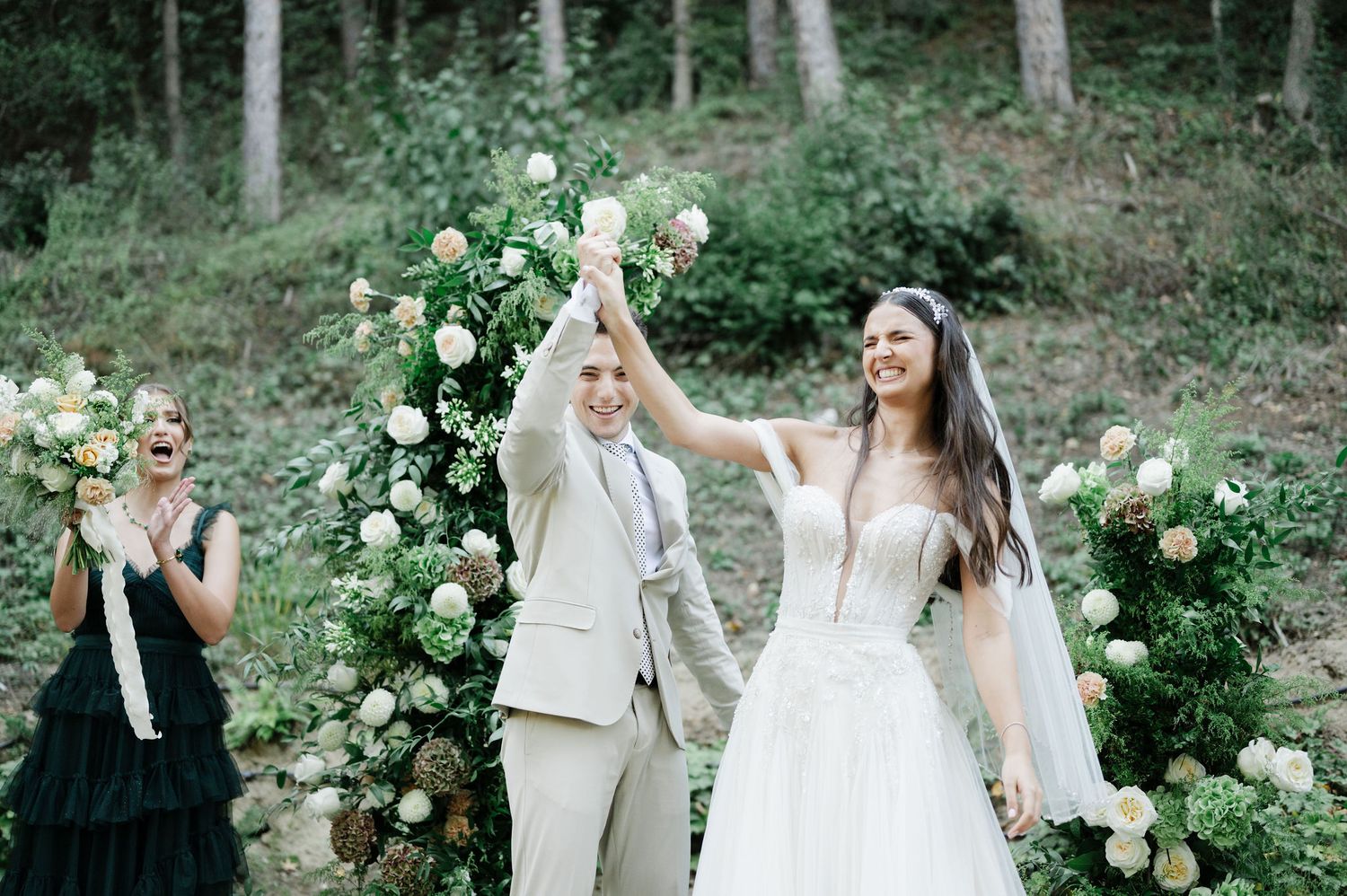 Newlyweds celebrate with raised arms under floral arch in woodland setting.