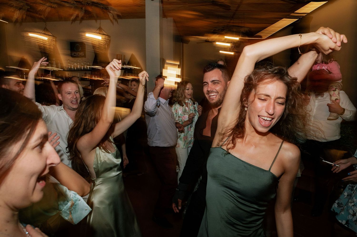 Wedding guests dancing and celebrating at a lively indoor reception party with warm lighting.