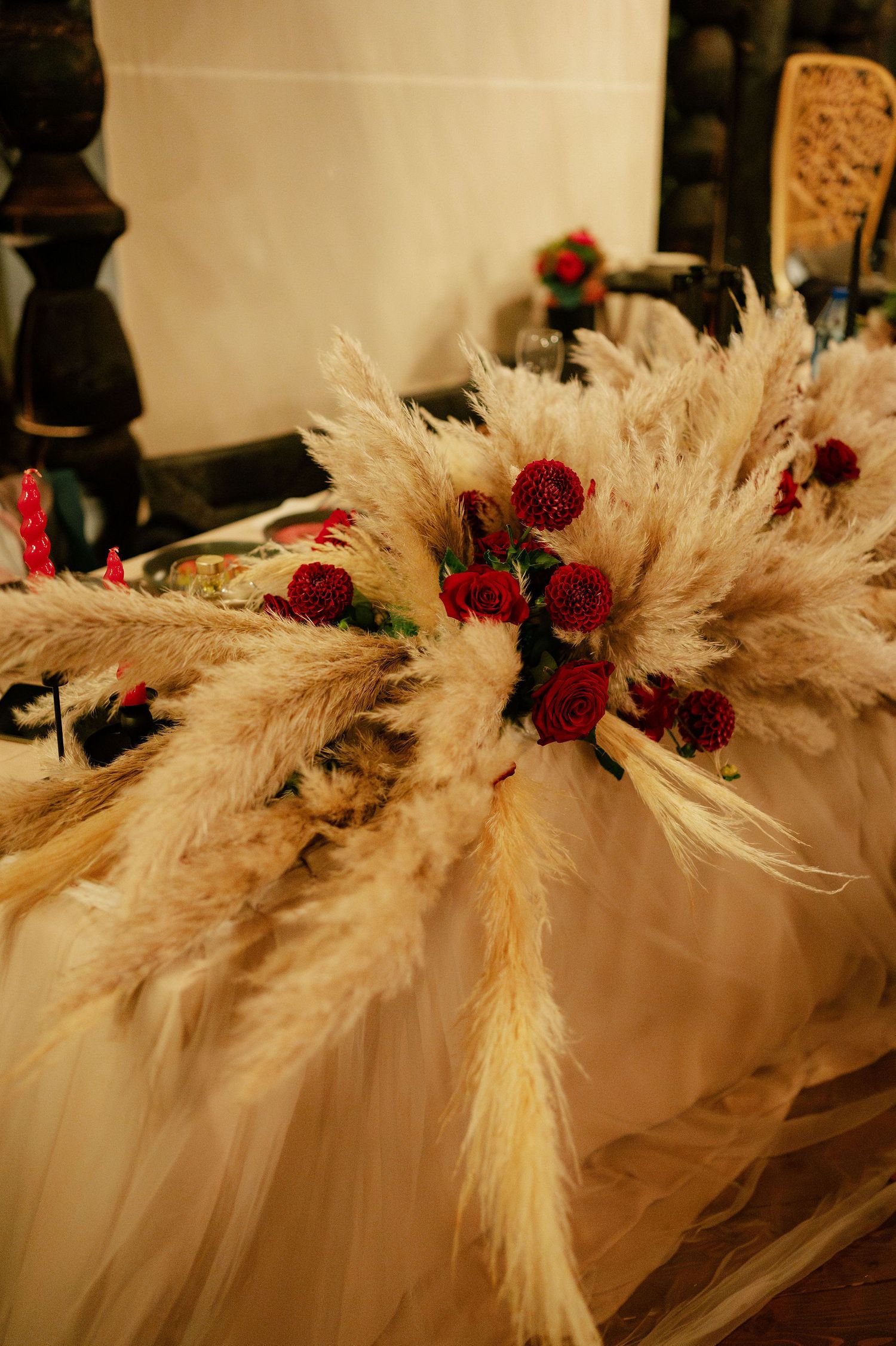Decorative arrangement of dried pampas grass with deep red flowers against white fabric.
