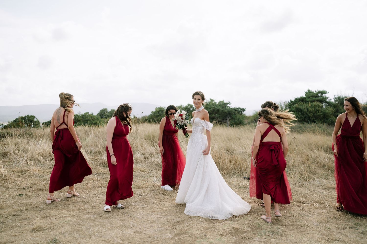 Wedding party in burgundy dresses and white gown pose together on a grassy hill.